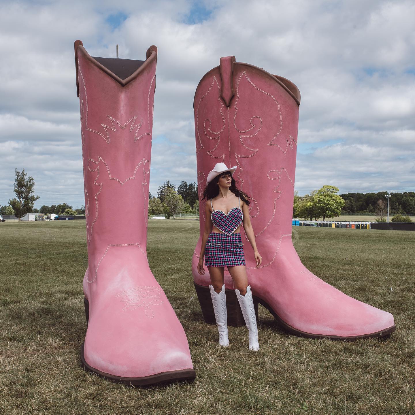 A pair of giant pink cowboy boots. In the middle is Briony Douglas wearing a dress, white cowboy hat and matching white cowboy boots.