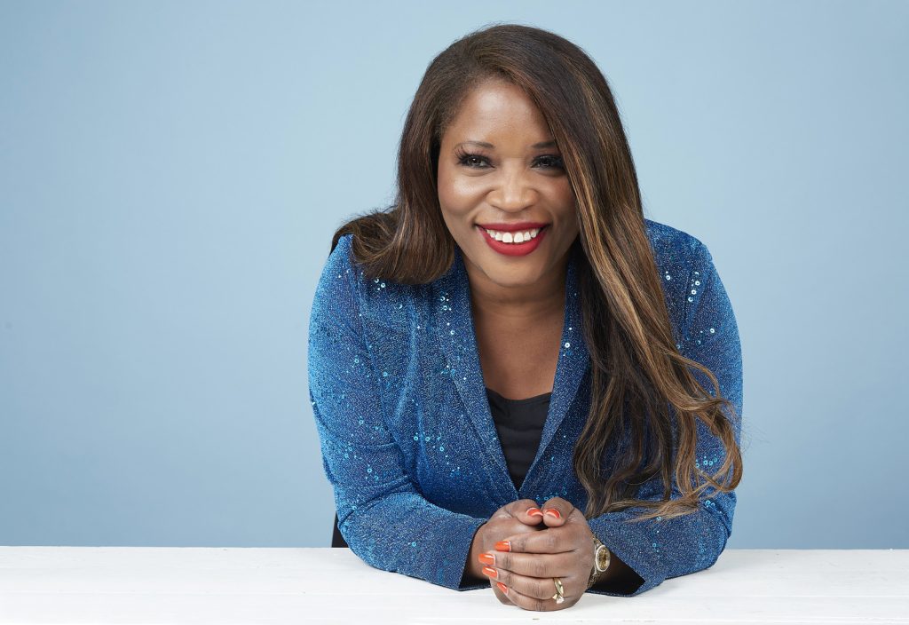 Claudette McGowan smiling wearing a sparkly blue blazer and leaning against a white tabletop. Her hands are closed and she is leaning slightly forward.