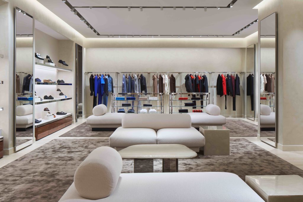 Long shot of the inside of a luxury clothing store. There is a rack of clothes on display, as well as a display of shoes on the side.