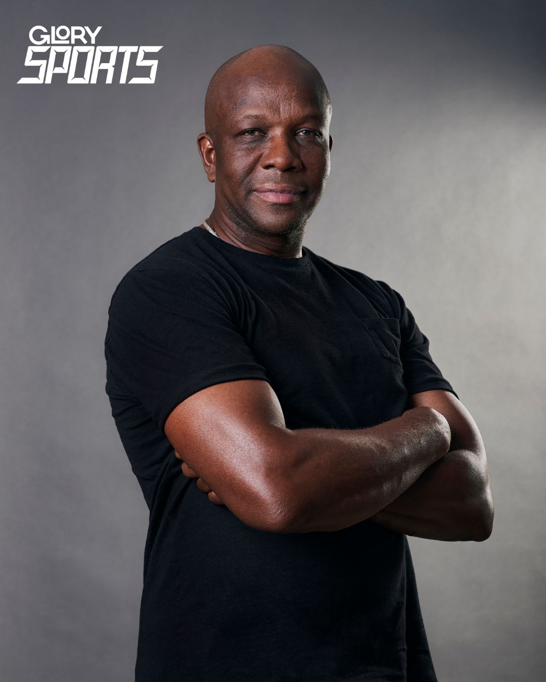 Olympian Donovan Bailey wearing a black t-shirt with arms crossed across his chest. He is looking directly into the camera against a grey background.
