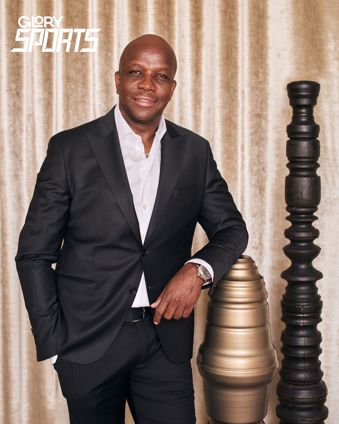 Donovan Bailey wearing a black suit and unbuttoned white dress shirt. He is smiling and leaning on a decorative plinth against gold curtains.