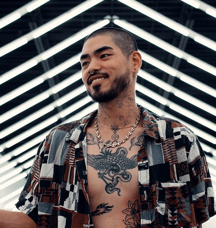 Man with his shirt open revealing a large chest tattoo of a bird eating a snake. There are lights behind the man in a chevron pattern.