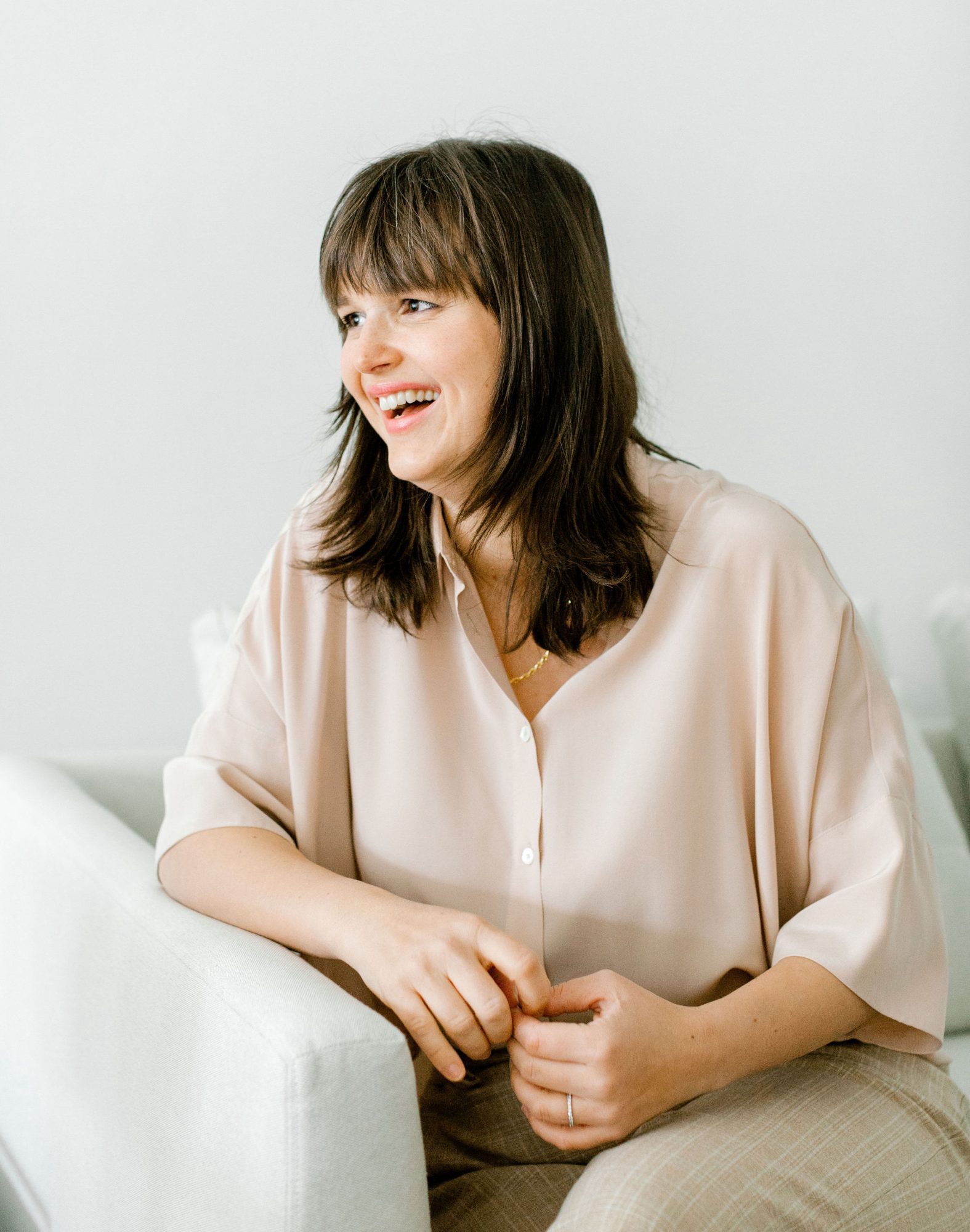 Barbora Samieian laughing and looking to the side wearing a blush pink blouse. She is leaning against a couch arm.