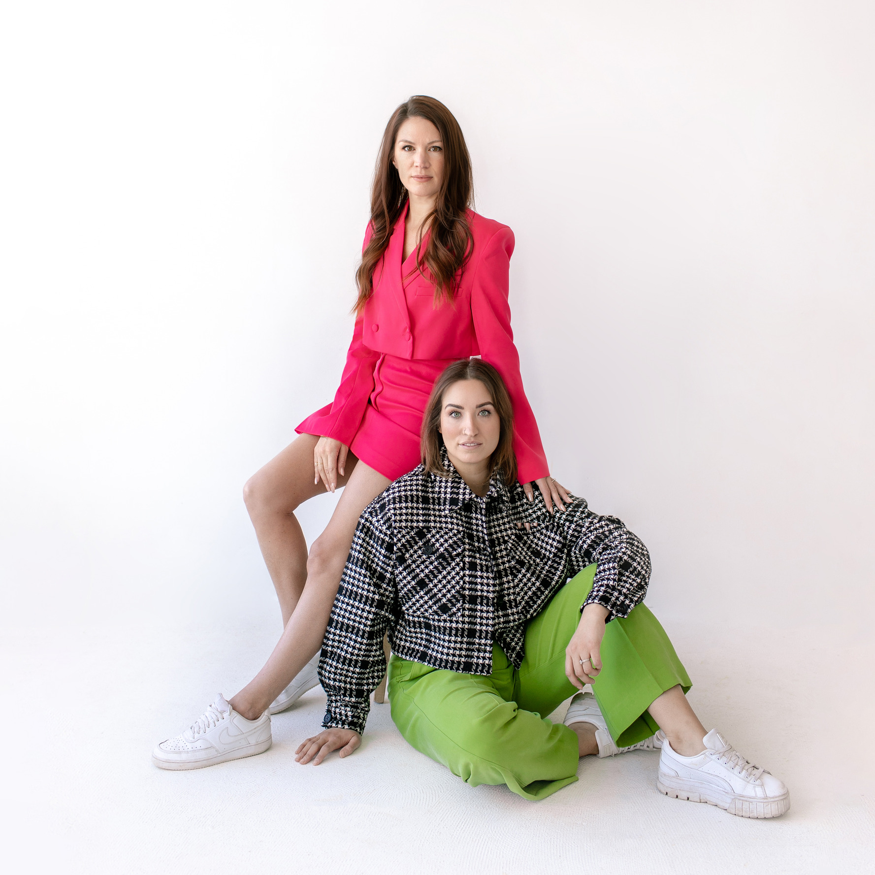 April Hicke seated on a stool wearing a fuschia pink blazer set and white sneakers. Marissa is seated on the ground wearing a plaid jacket and green pants.