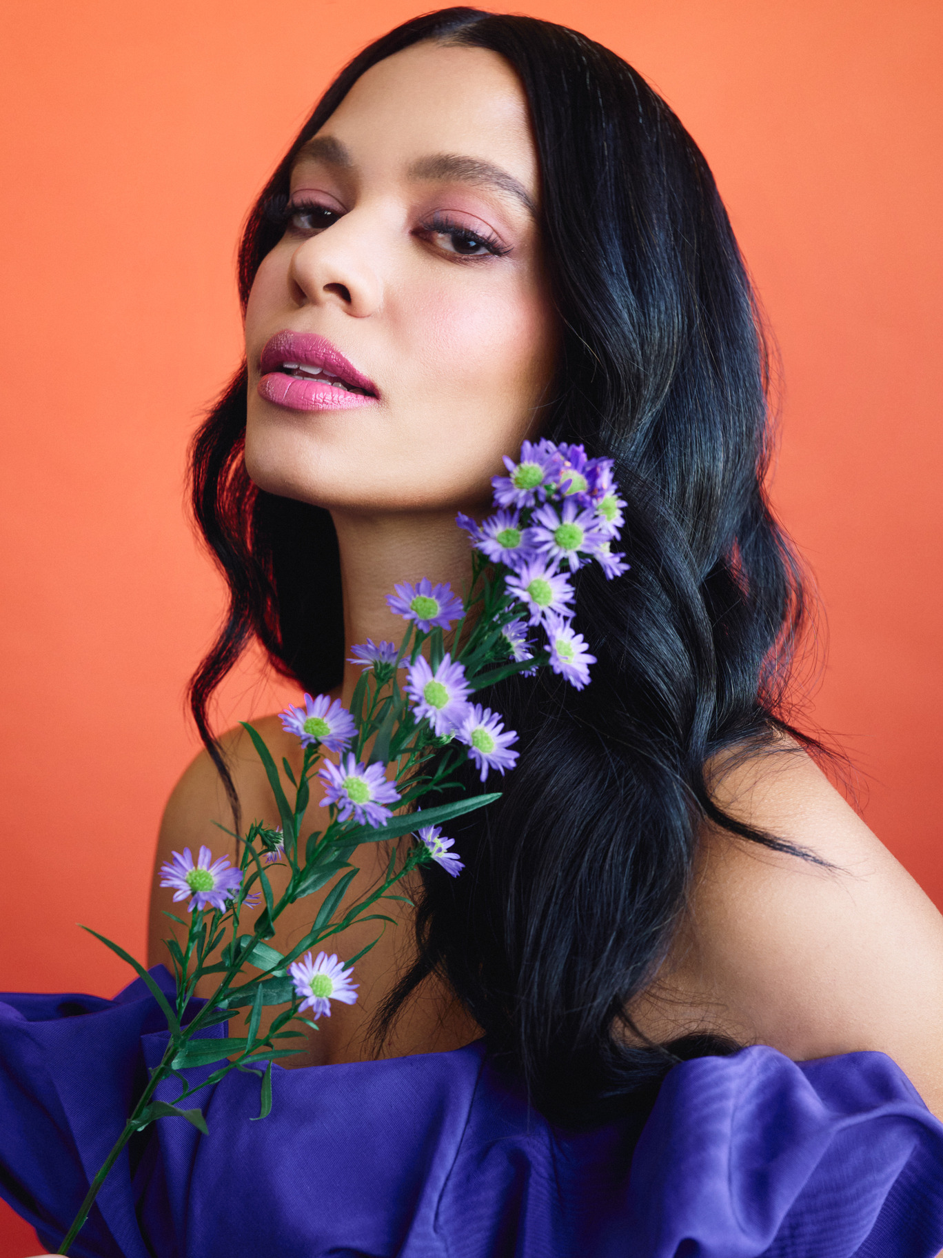 Aurora James is wearing an off the shoulder violet dress. Her hair is down and head is tilted looking into the camera. A bushel of lavender paper flowers frames her face. She is against an orange background.