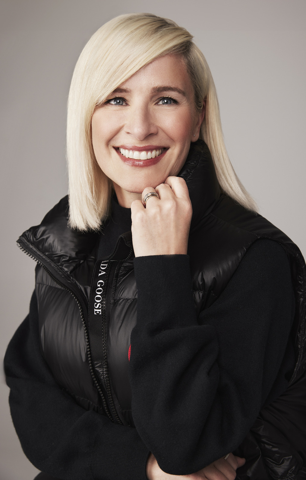Carrie Baker wearing a black Canada Goose puffer vest and black long-sleeve sweater underneath. She is smiling with blonde hair perched on her hand.