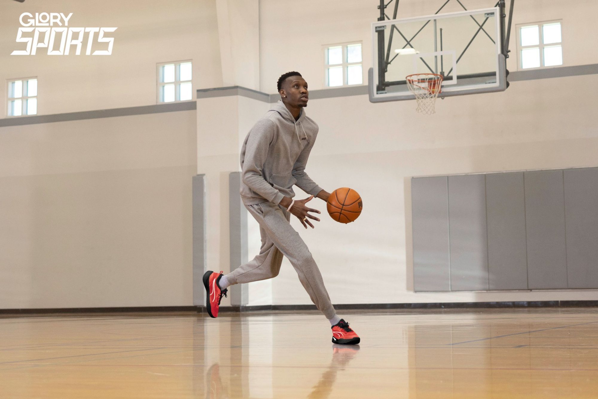Chris Boucher wearing a grey sweatsuit and red basketball shoes. He is dribbling a basketball on a court.