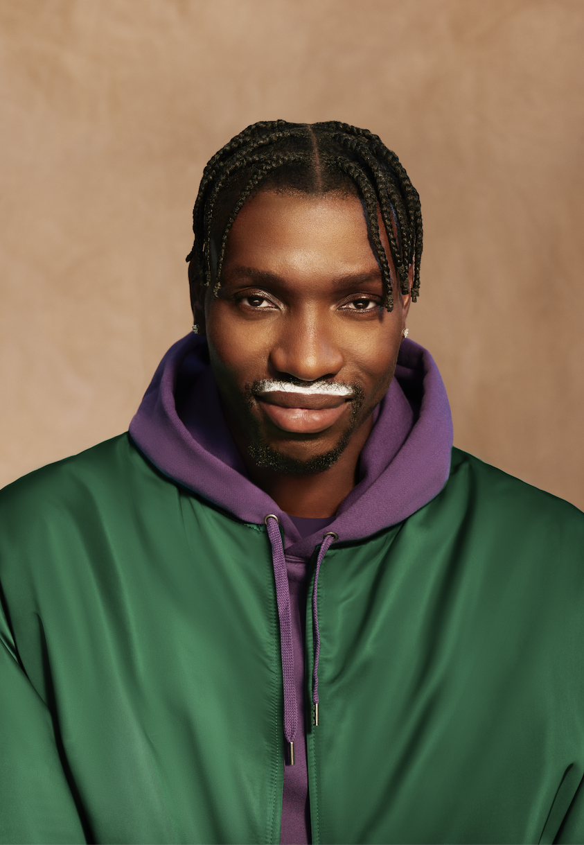 Chris Boucher wearing a green jacket and a purple hoodie underneath. He is against a brown backdrop and staring in the camera with a milk moustache