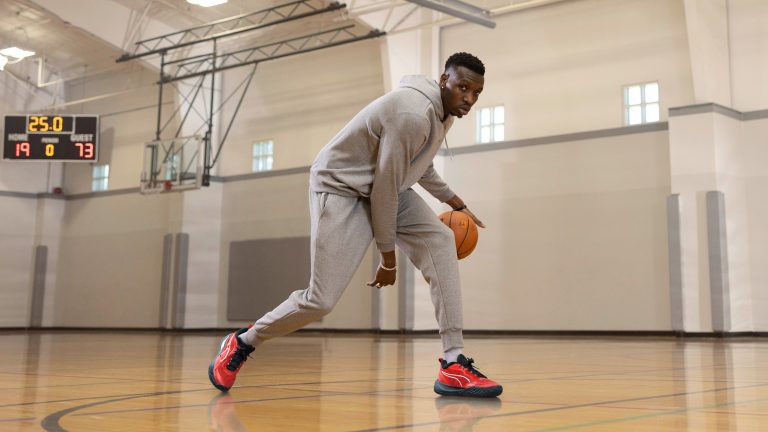 Chris Boucher wearing a grey sweatsuit and red basketball shoes. He is dribbling a basketball on a court.