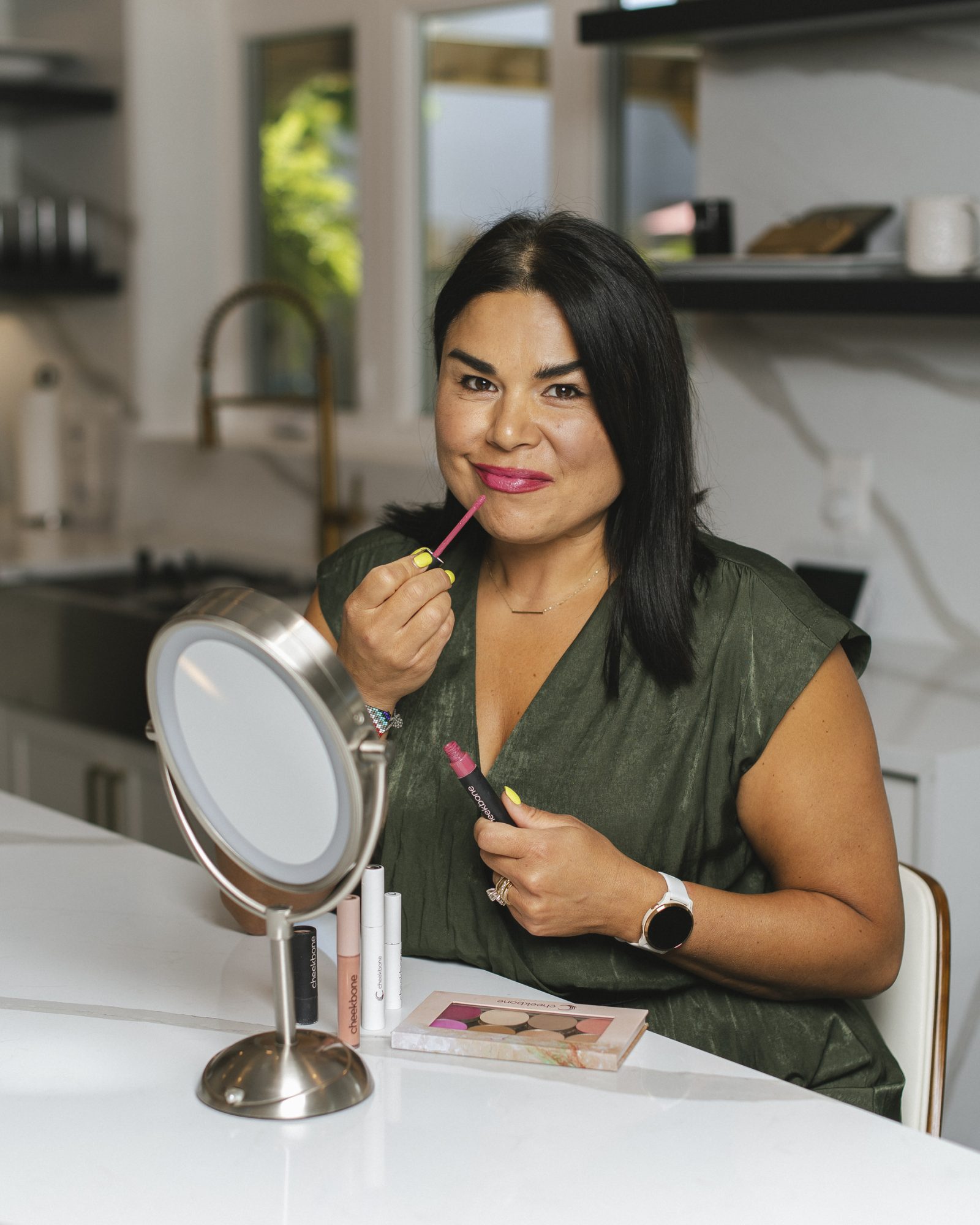 Jenn Harper wearing an olive green blouse. She is seated at a marble counter with a makeup mirror and is applying lip gloss in a home kitchen.