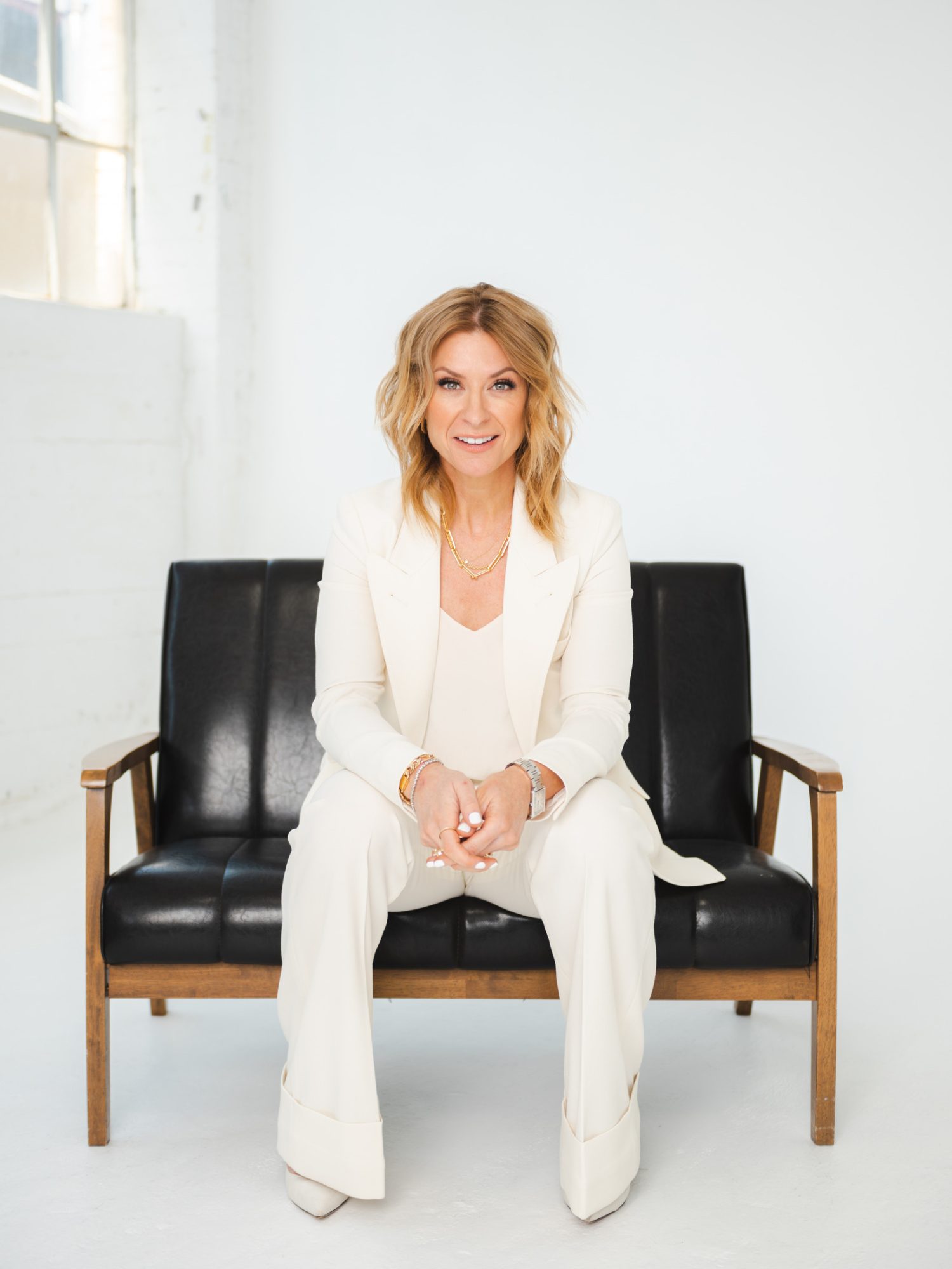 Jennifer Flanagan wearing a white suit. She has tussled wavy blonde hair and is seated on a leather loveseat in a white room.