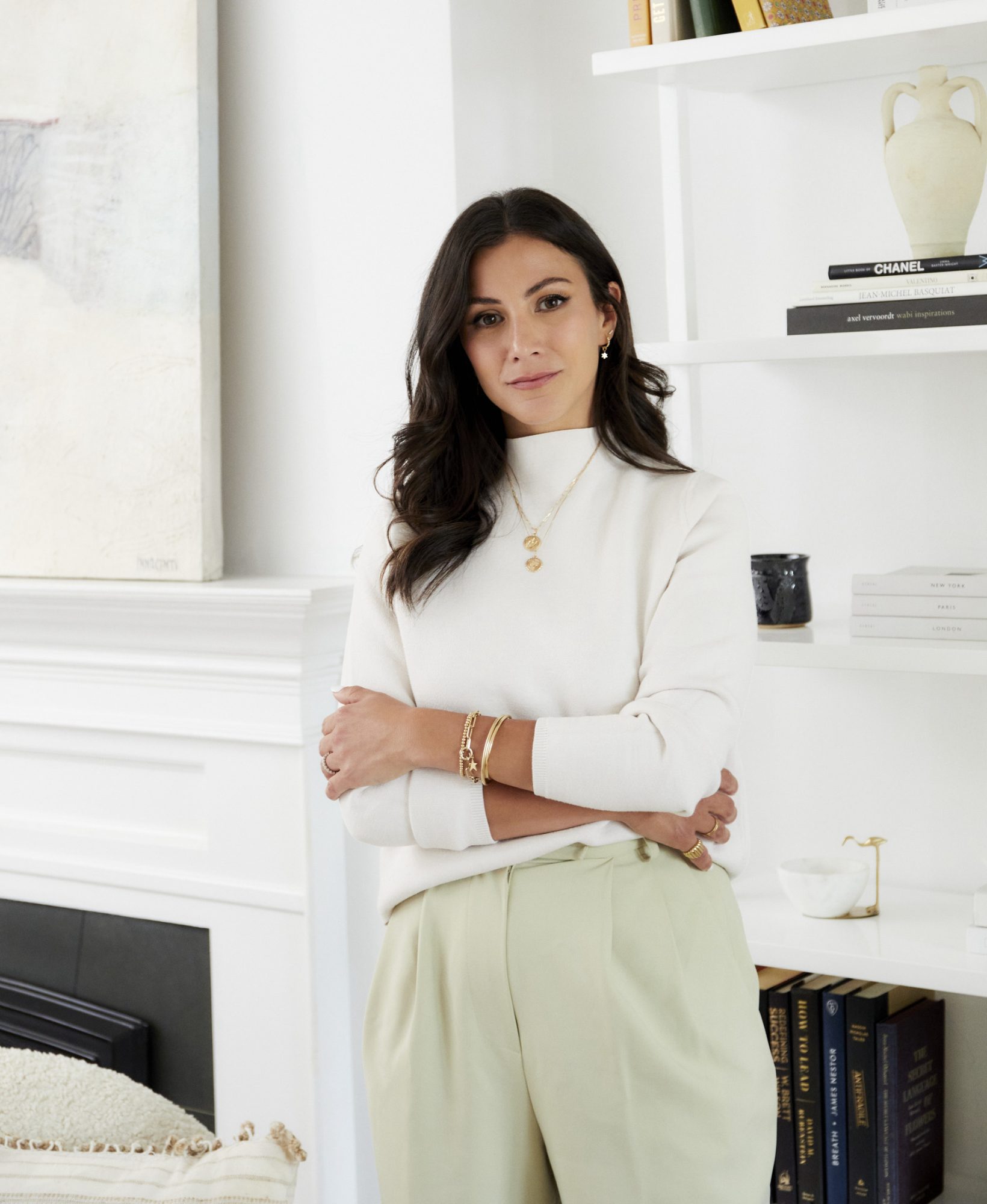Noura Sakkijha wearing a white turtleneck sweater with her arms crossed and white trousers. She is wearing Mejuri jewelry standing in a living room.