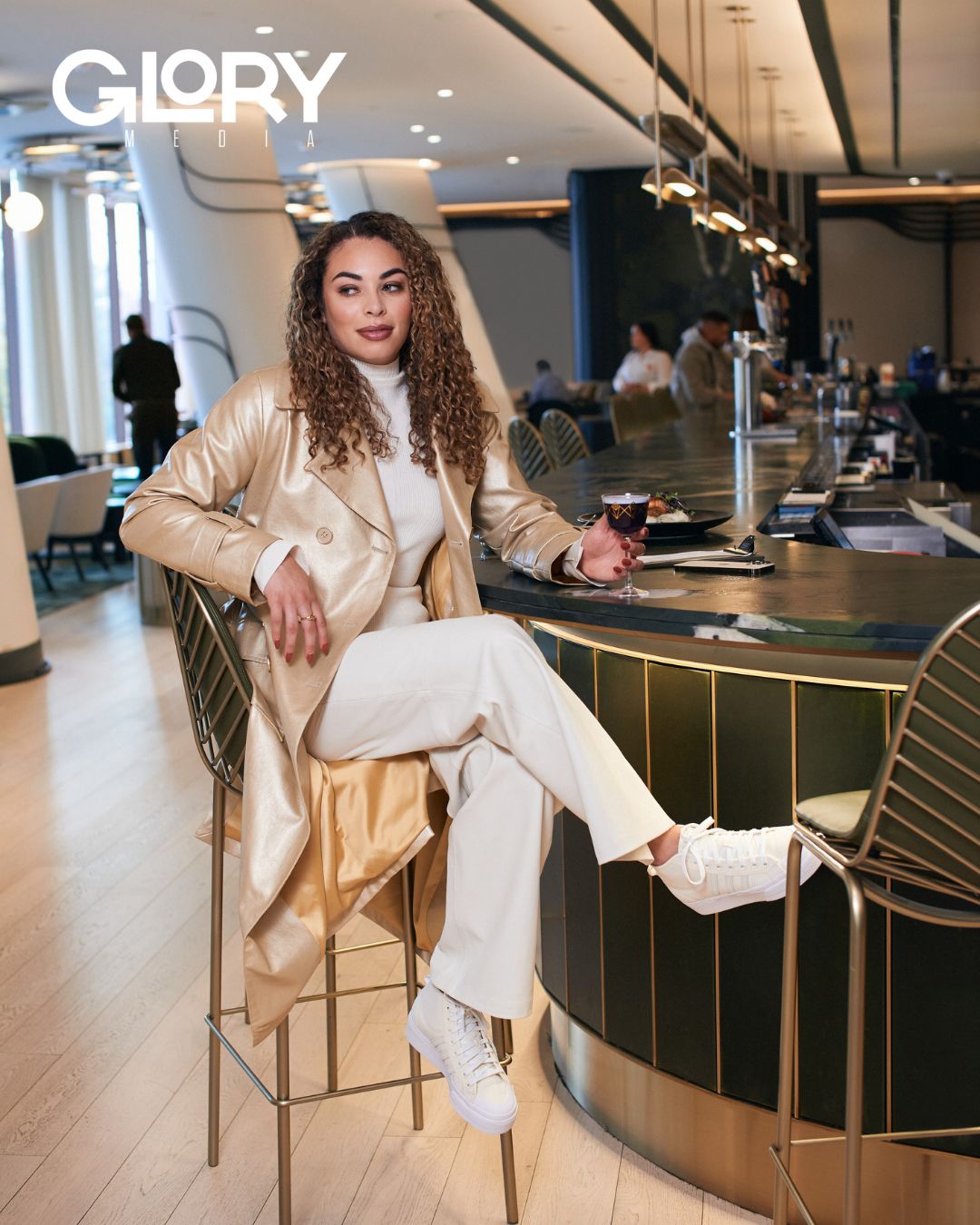 Sarah Nurse wearing a white turtleneck, a gold jacket, and pants seated at a hotel bar.