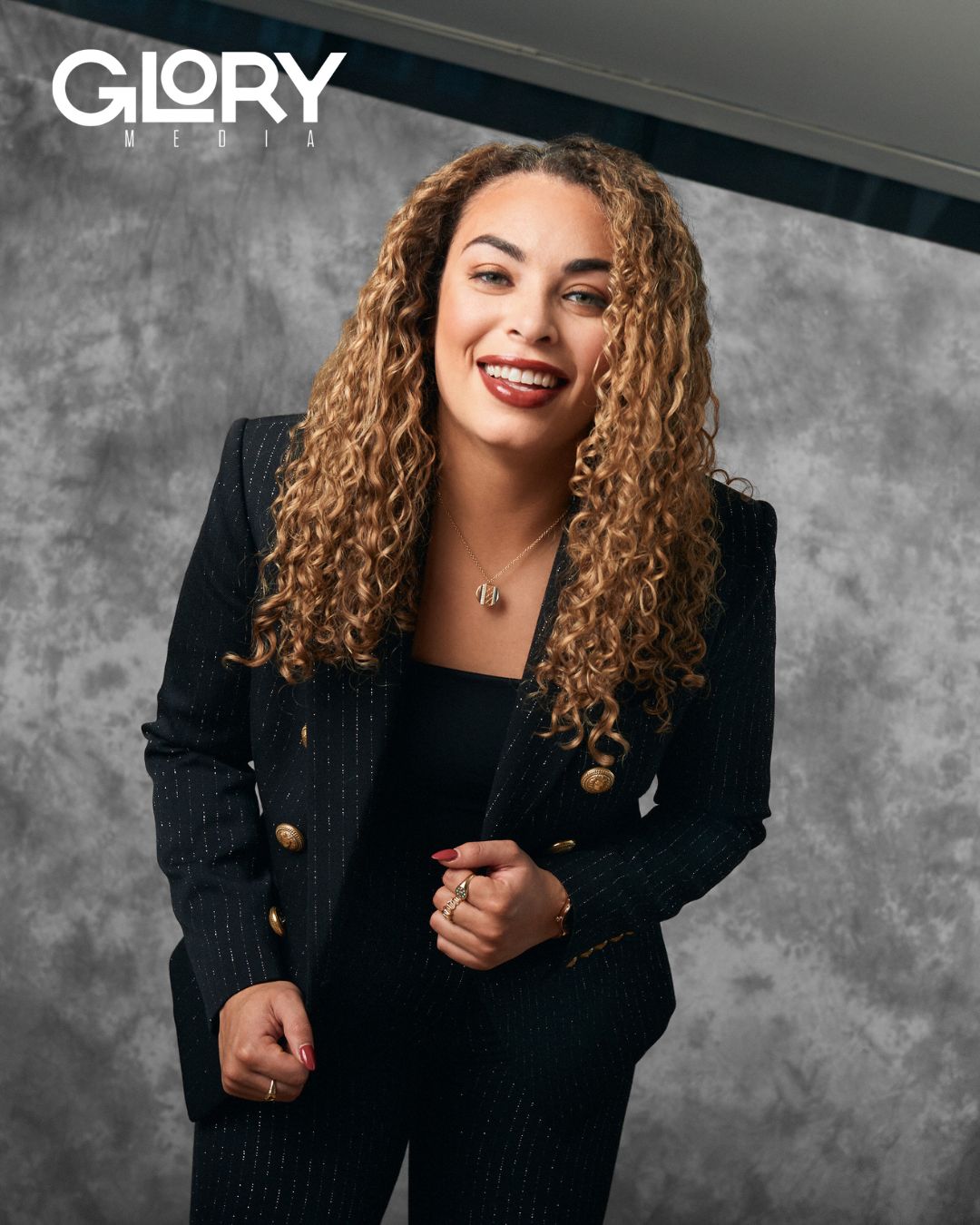 Sarah Nurse smiling into the camera. She is wearing a black suit against a grey background.