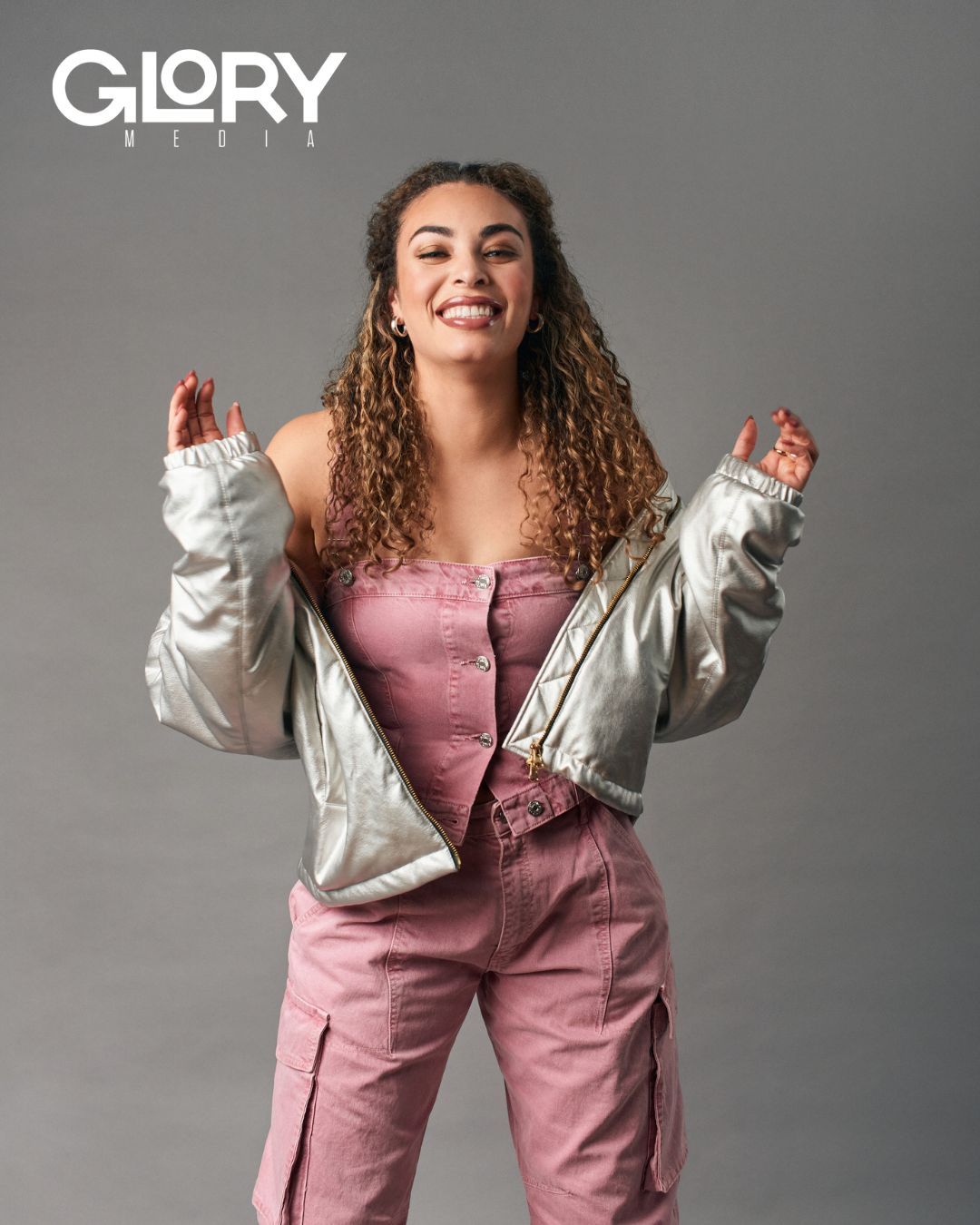 Sarah Nurse wearing a silver puffer and a pink jumpsuit against a grey background.