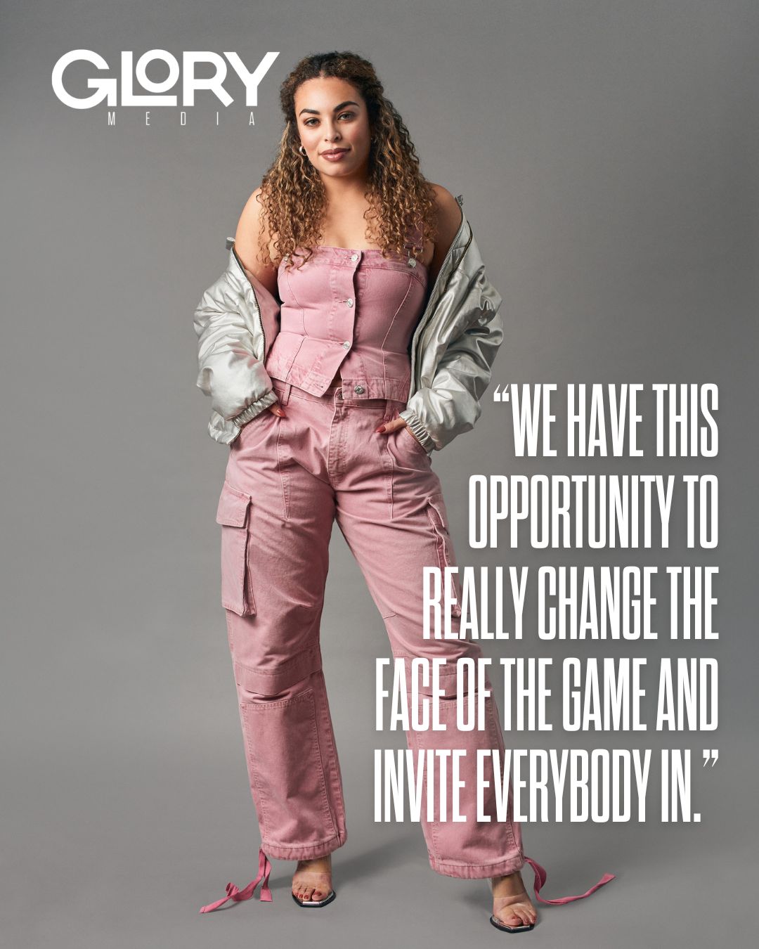 Sarah Nurse wearing a silver puffer and a pink jumpsuit against a grey background.