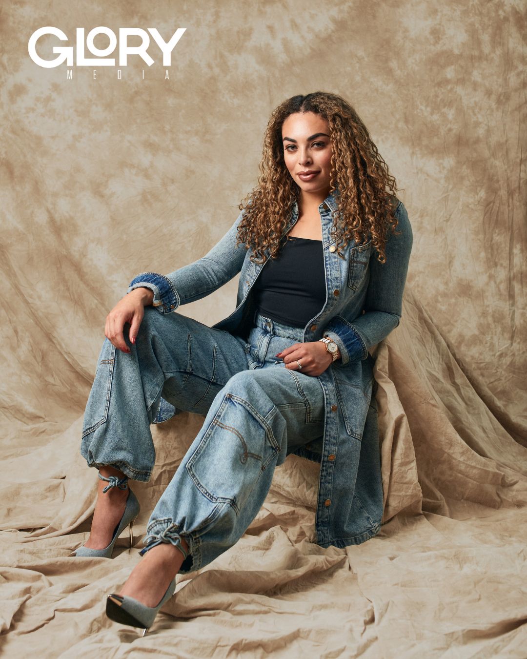 Sarah Nurse reclined on a draped seat. She is wearing a denim jacket and pants with matching shoes.