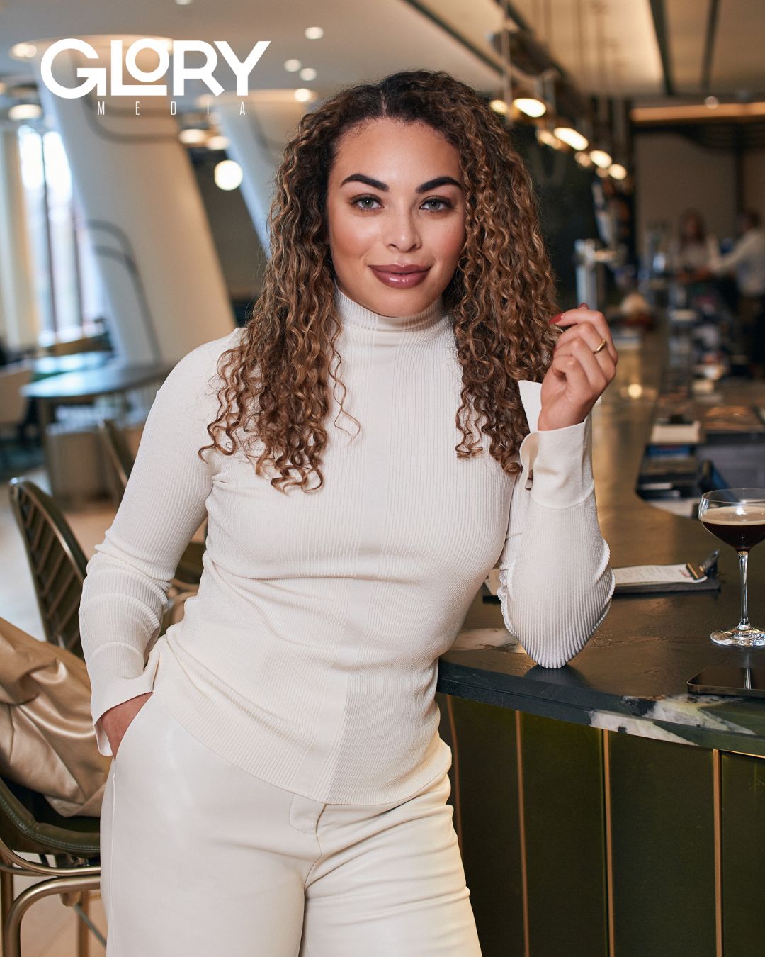 Sarah Nurse wearing a white turtleneck and pants leaning against a hotel bar.