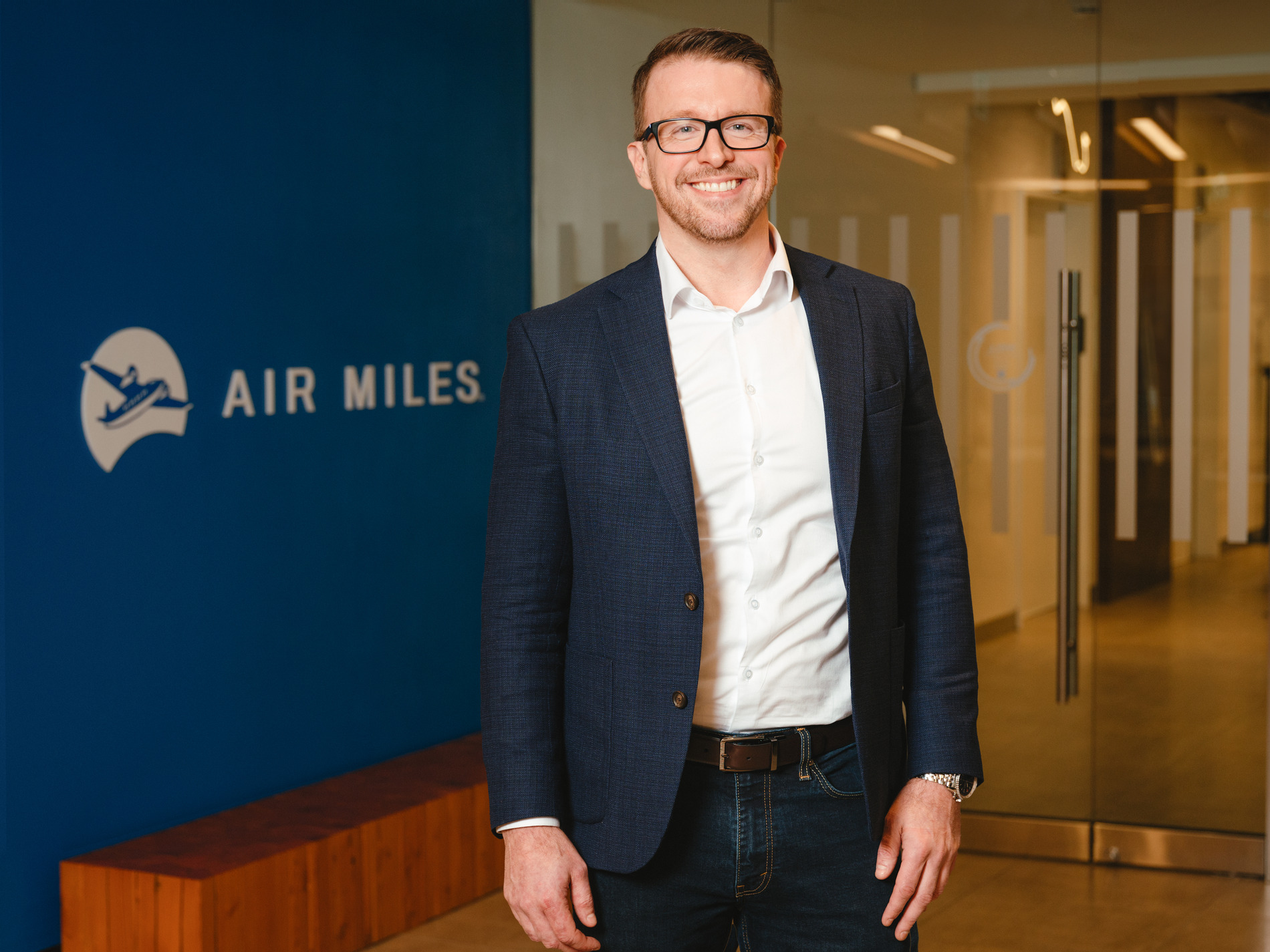 Shawn Stewart in a white button up shirt and navy jacket and jeans standing and smiling in front of an AIR MILES sign.