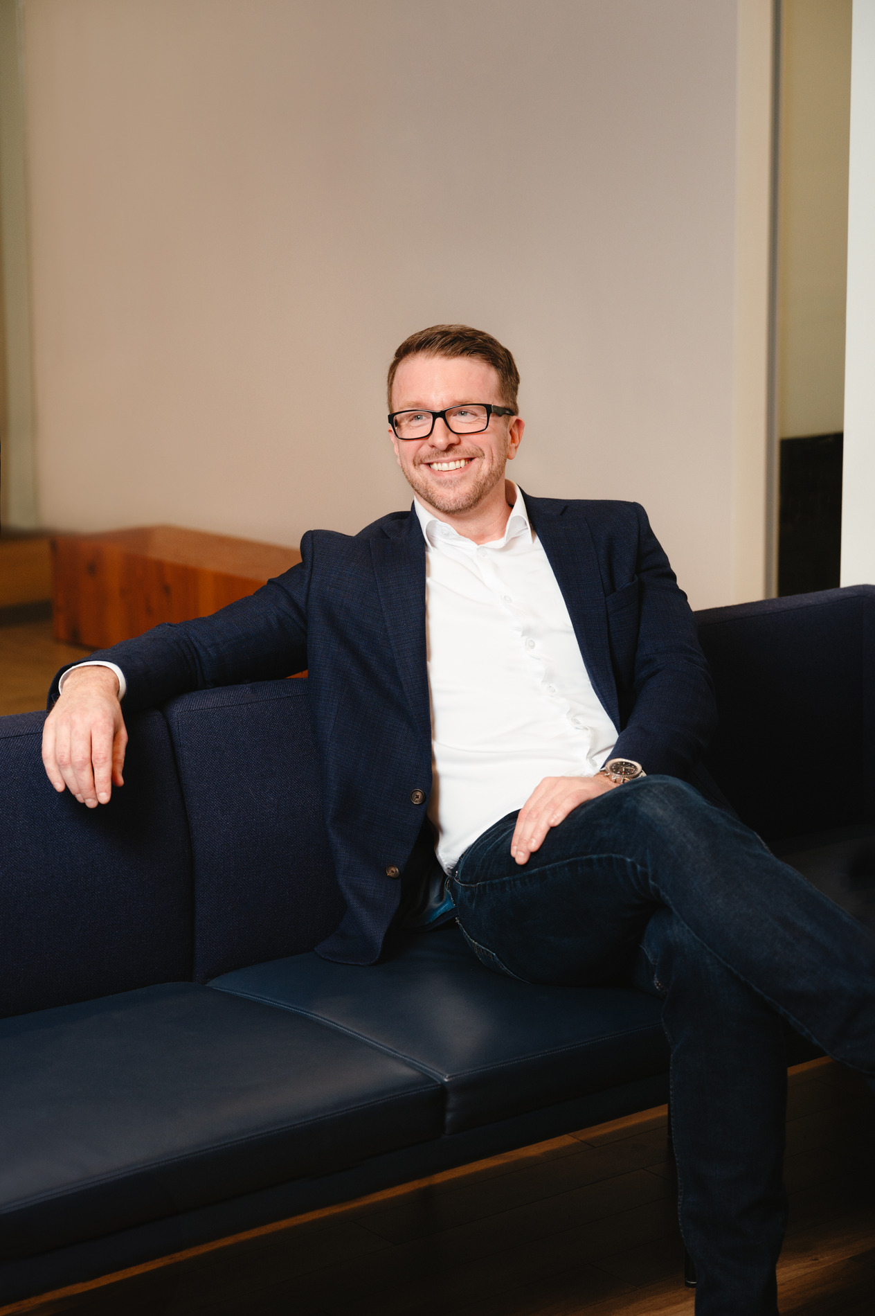 Shawn Stewart in a white button up shirt and navy jacket and jeans reclining on a couch and smiling off to the side of the camera.