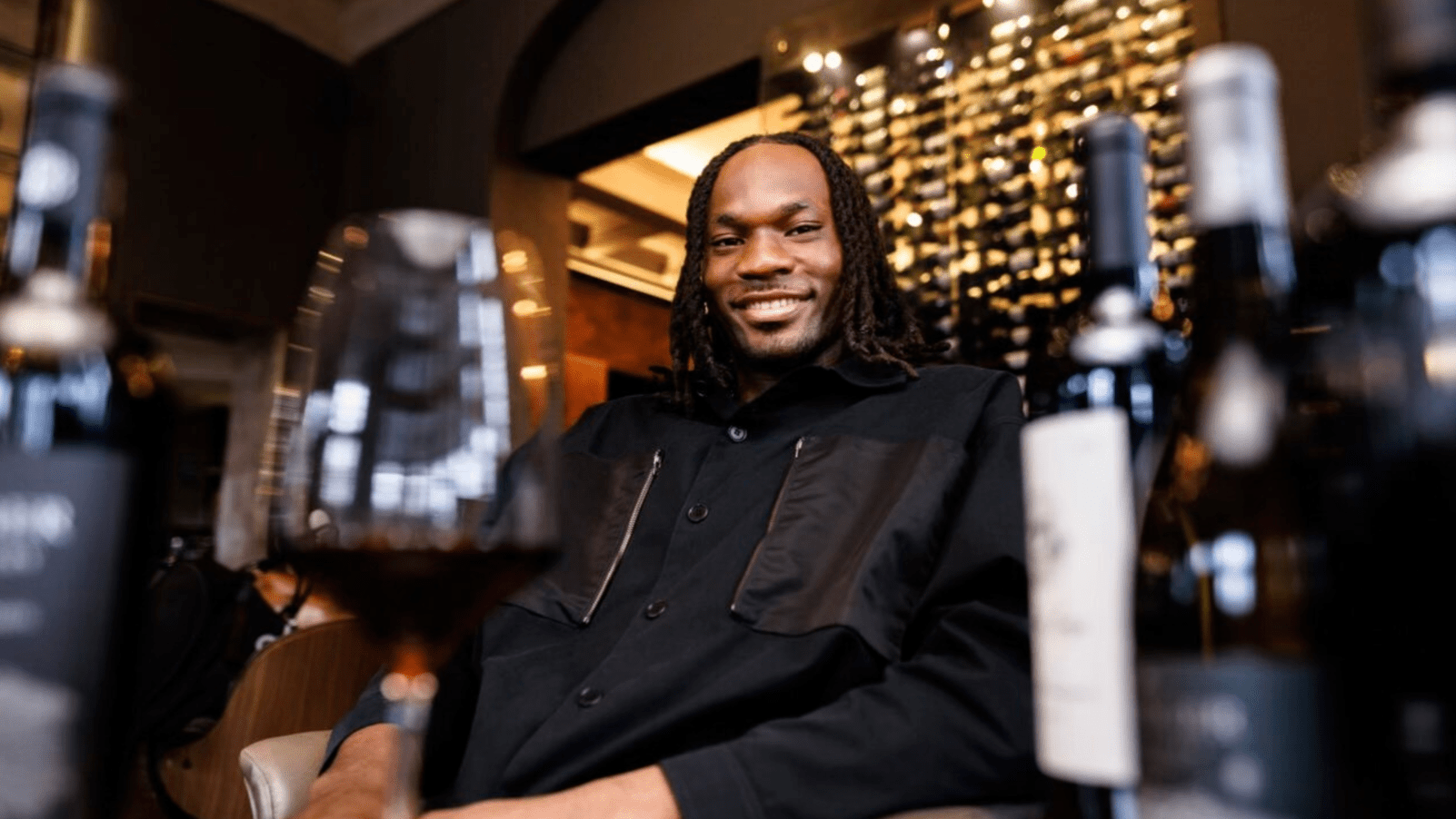 Precious Achiuwa smiling and seated inside a wine cellar with a glass of red wine