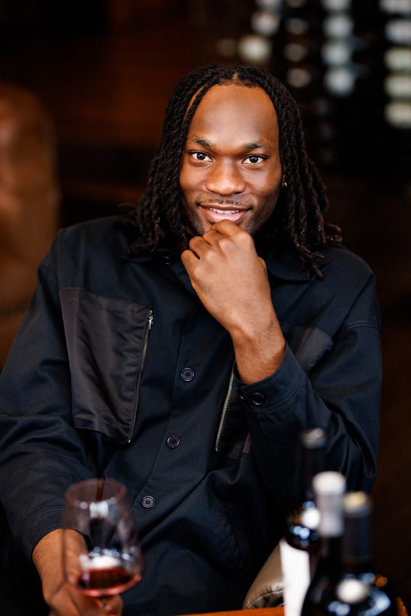 Precious Achiuwa dressed in black with a smirk, resting his chin in his hand while holding a glass of red wine.