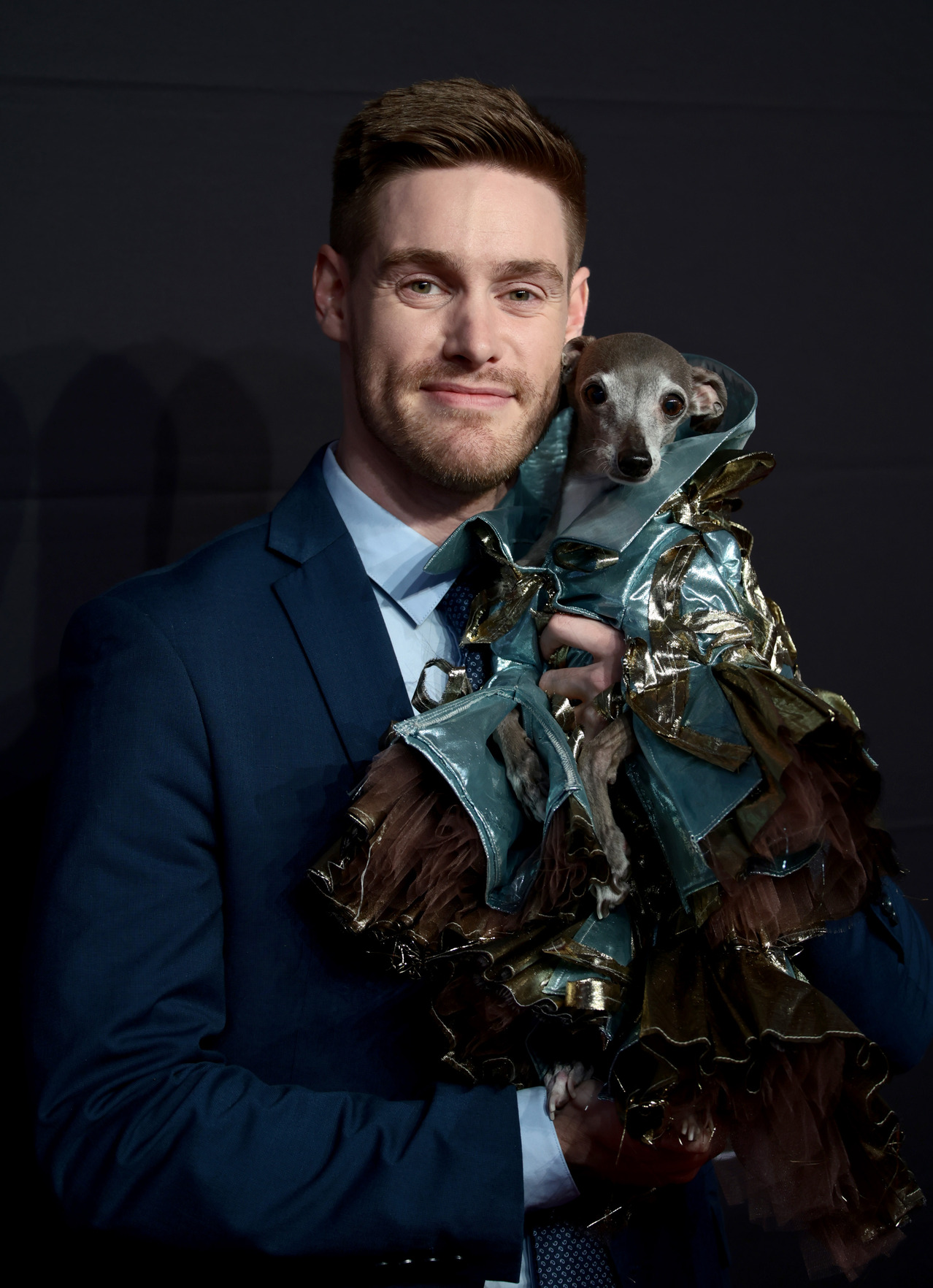 Thomas Shapiro in a blue suit and shirt is holding Italian greyhound Tika the Iggy, who is wearing a shiny raincoat.