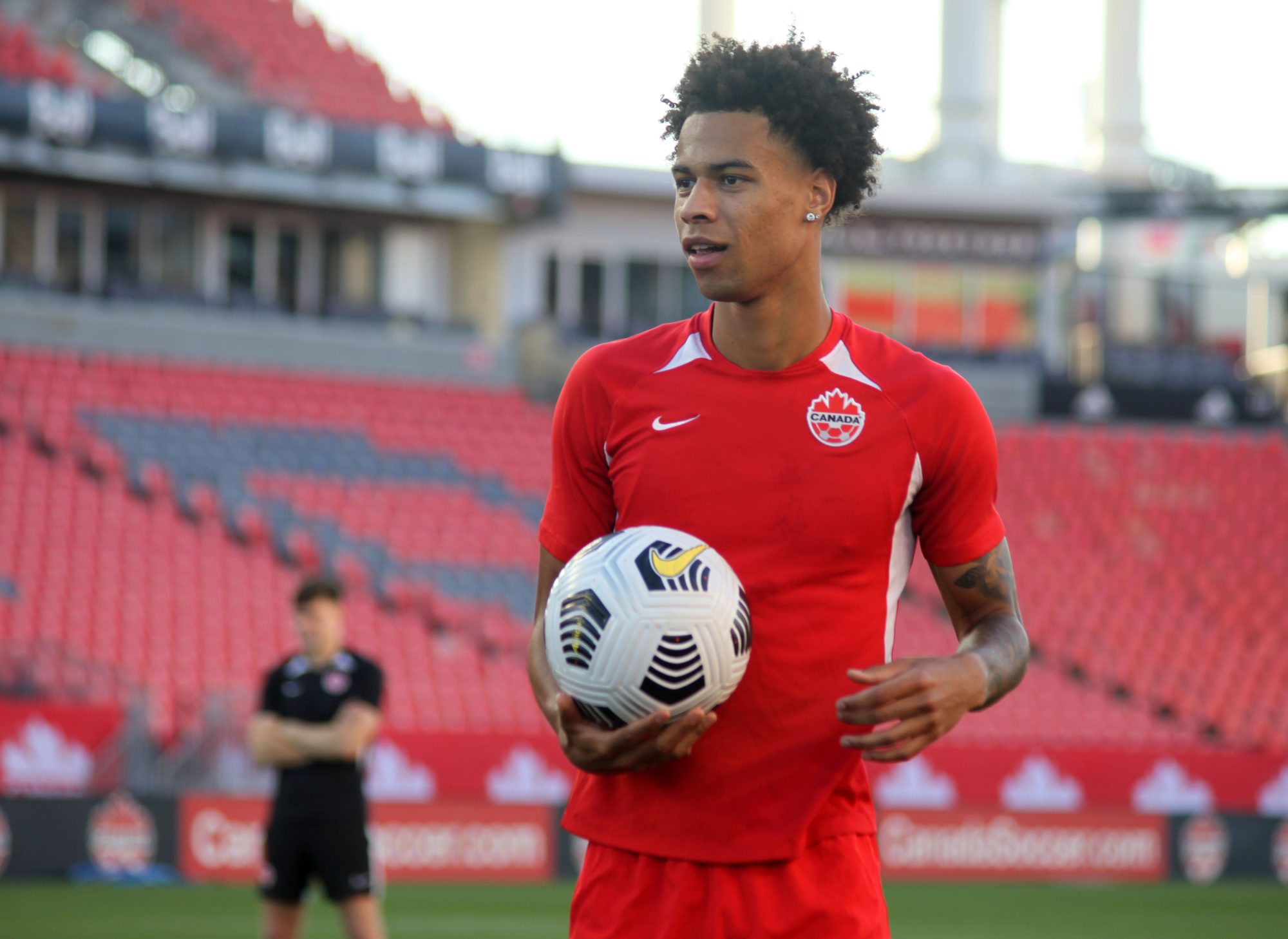 Tajon Buchanan in a red jersey holding a soccer ball against the backdrop of a stadium with red seats.
