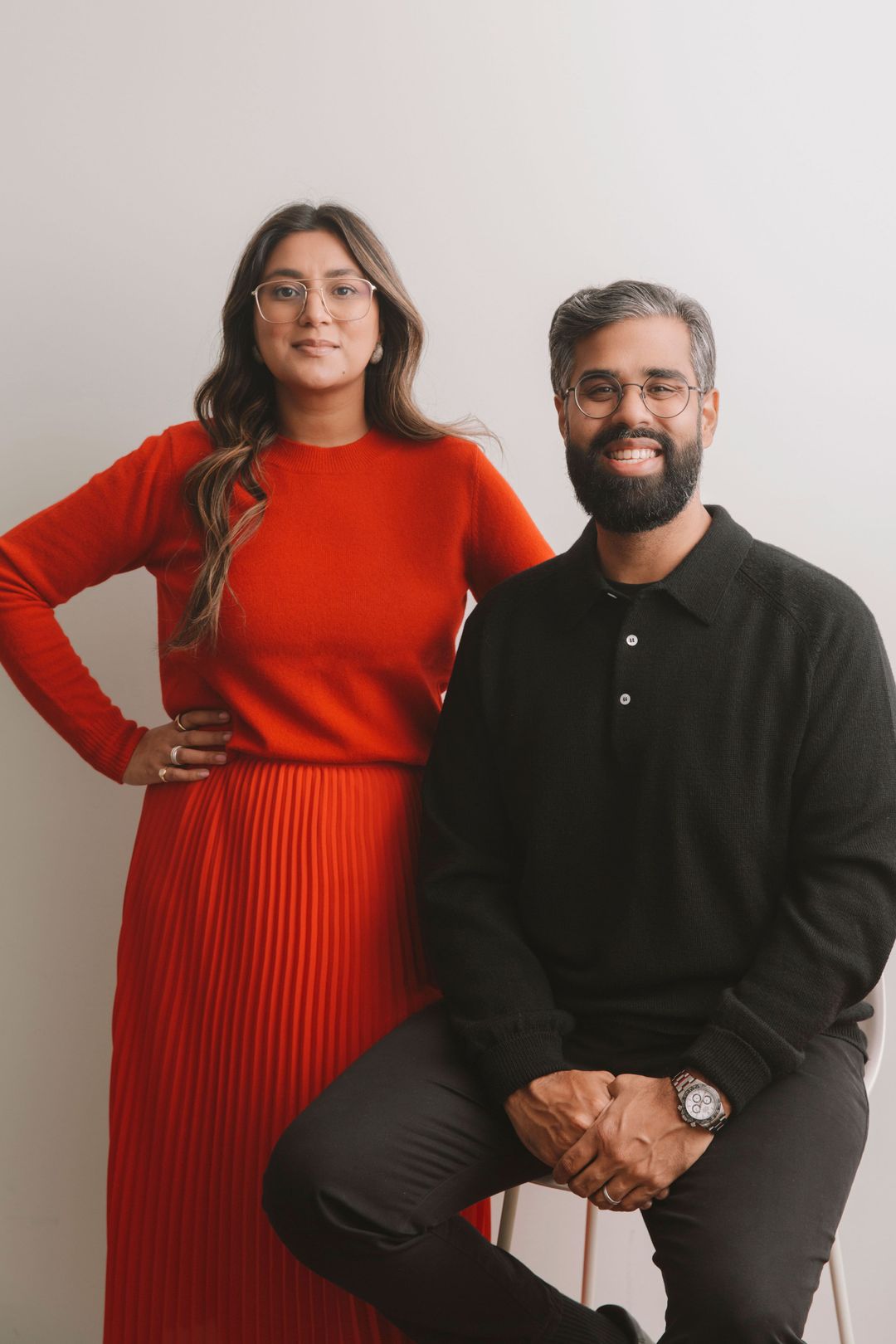Arati Sharma standing with her hair down and wearing a red sweater and dress. She is standing behind Satish Kanwar, who is sitting on a stool and dressed all in black.