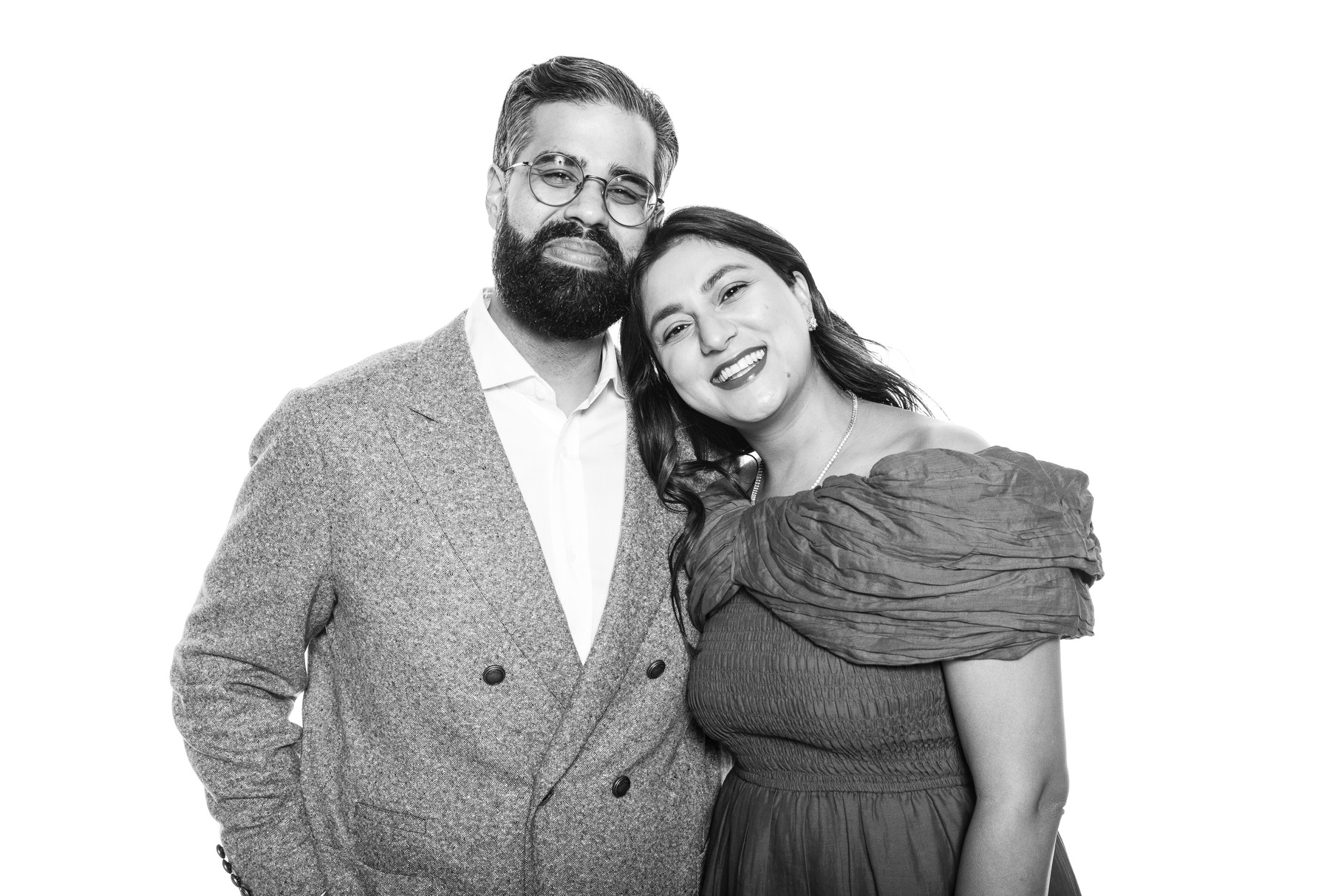 Black and white photo of Satish Kanwar and Arati Sharma. He is wearing a double breasted jacket with a white button up shirt. She is wearing a festive dress. They are both smiling and leaning against each other.