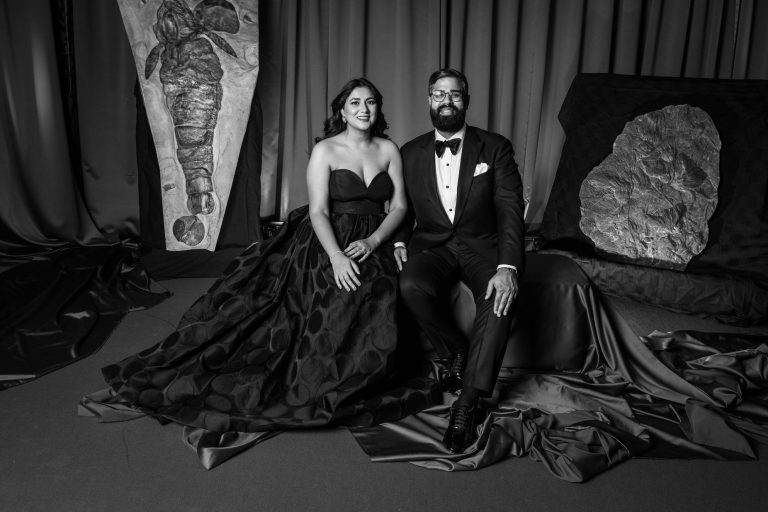 Black and white photo of Arati Sharmi seated next to Satish Kanwar for a portrait session. She is dressed in a voluminous ball gown and he is in a tuxedo.
