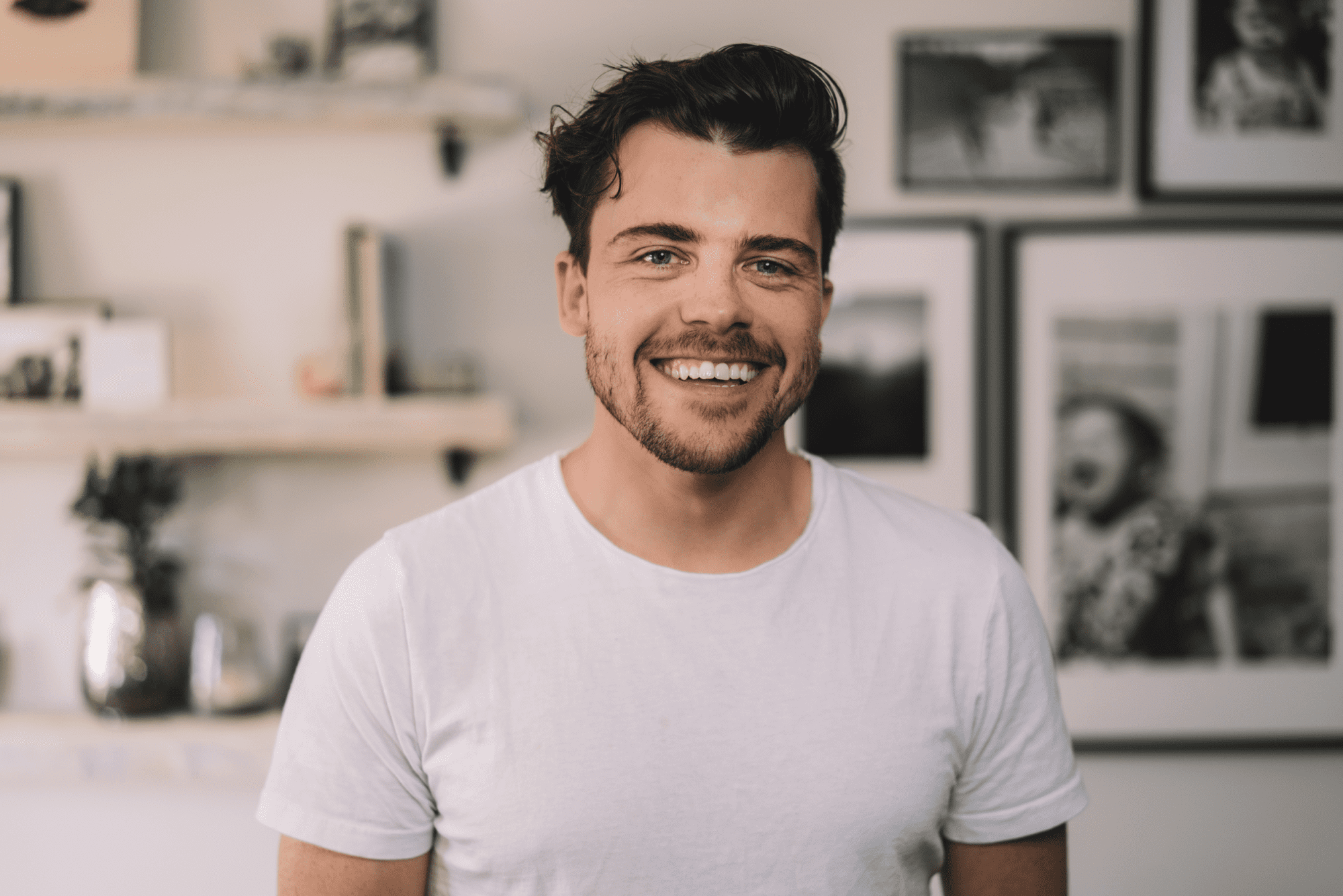 Daniel Eberhard smiling with wavy brown hair. He is wearing a white t-shirt and in the background are framed photos.