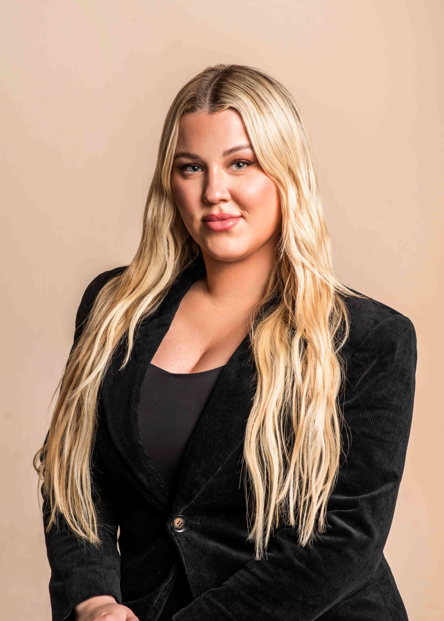Mallory Greene headshot with long blonde hair against a beige backdrop. She is wearing a black blazer and shirt.