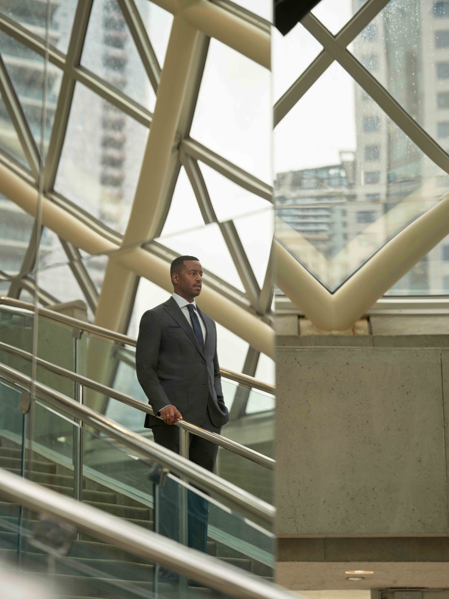 Toronto Symphony Orchestra CEO Mark Williams wearing a charcoal grey suit and tie. He is on a stairwell with his hand grasping the metal railing with the facade of the glass window behind him.