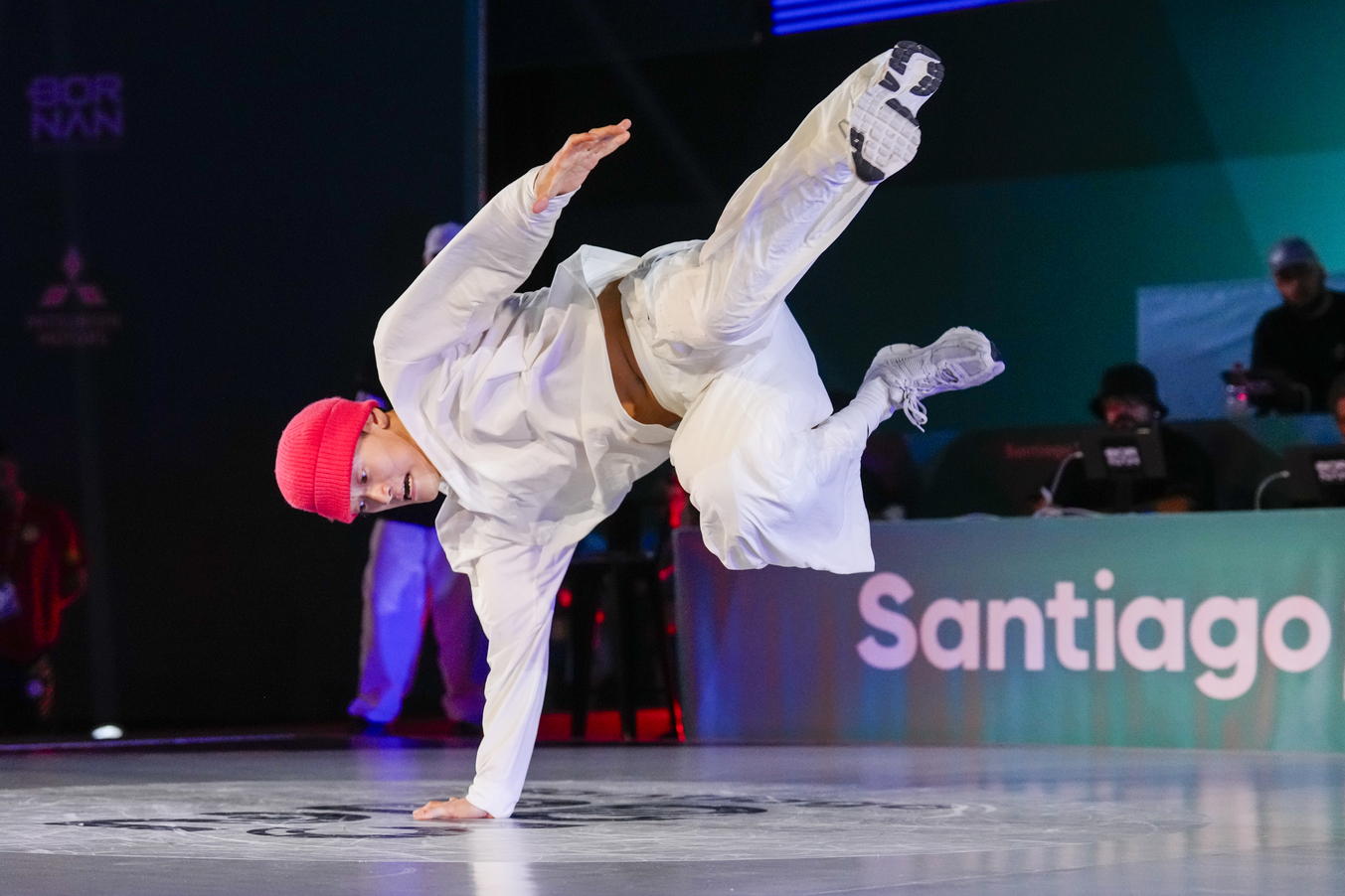 Breakdancer Phil Kim in a white outfit and pink hat performing a break move on the dancefloor at a competition.