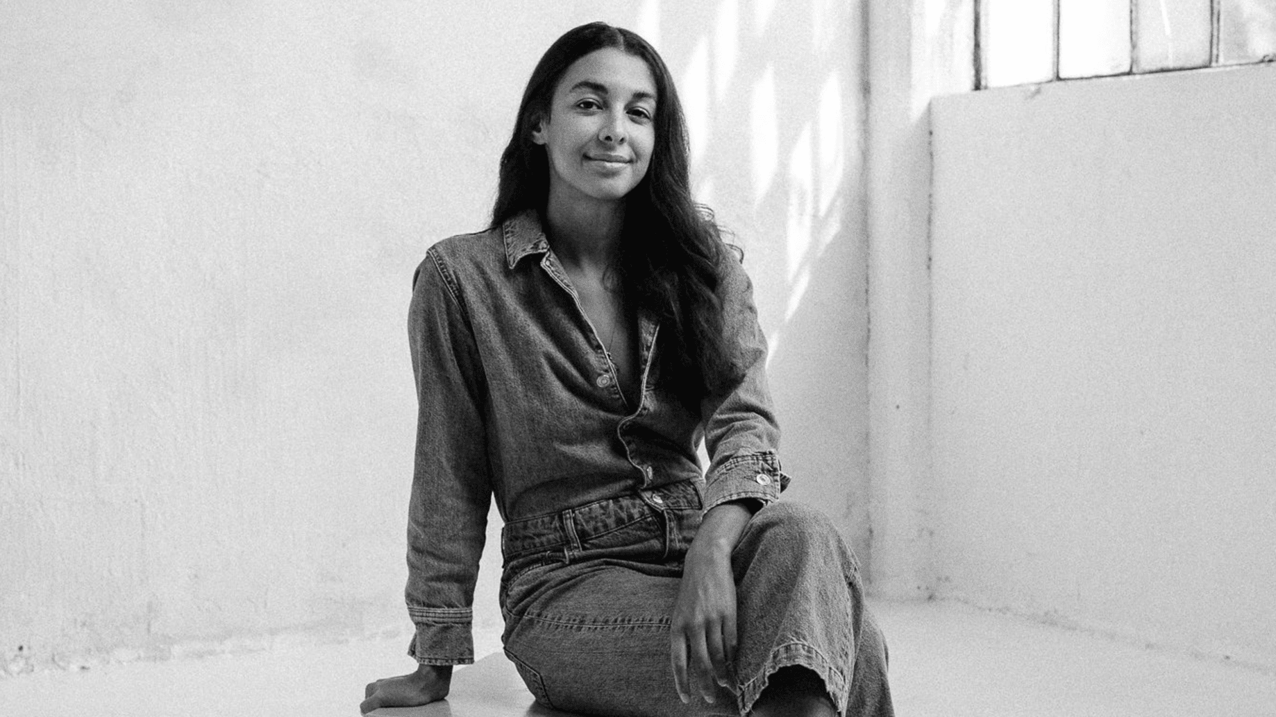 Black and white image of Daphné Eastmond in a denim shirt and jeans sitting on a stool and looking into the camera.