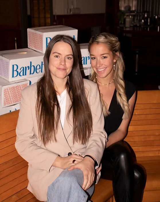 Andrea Grand and Katie Fielding sitting on a wooden bench and a box of Barbet behind them. They are smiling into the camera.