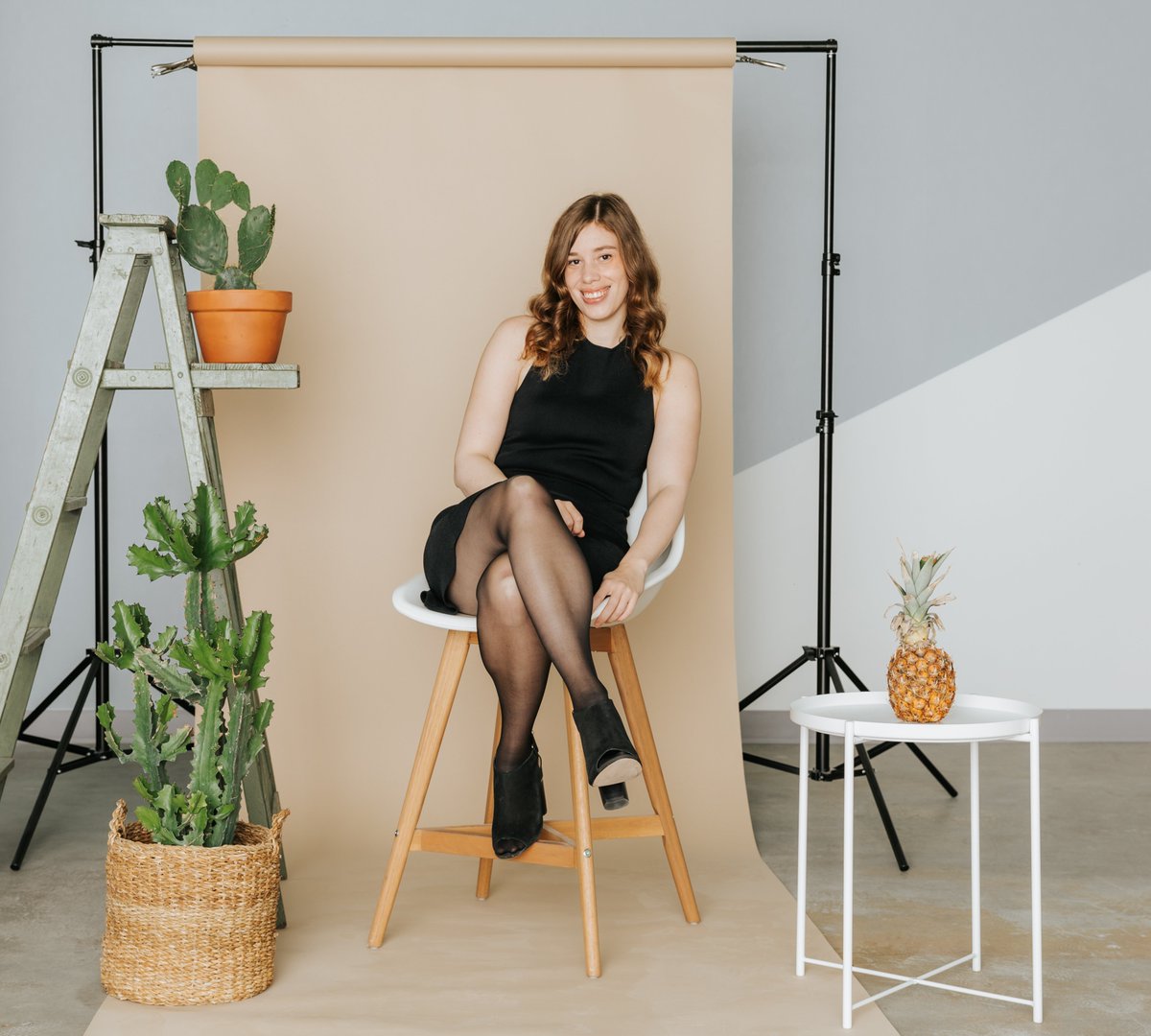 Katherine Homuth sitting in a black dress and Sheertex stockings on a stool set against a backdrop with cacti surrounding her.