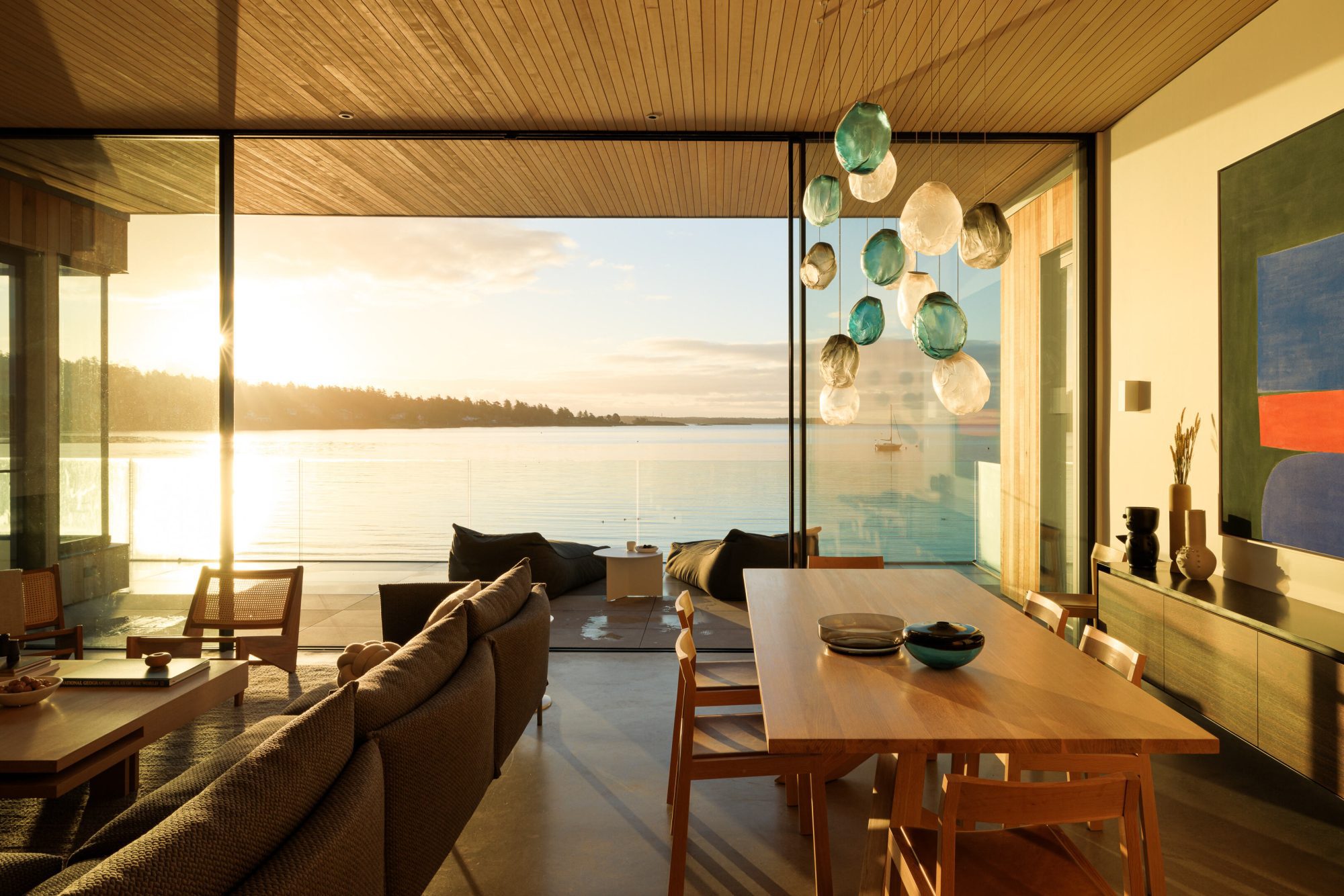 Sun-filled living and dining room looking out on an oceanscape with a tree-covered mountain in the distance. There is a water-glass chandelier hanging overtop a light brown dining table and chairs with large sliding patio doors open.