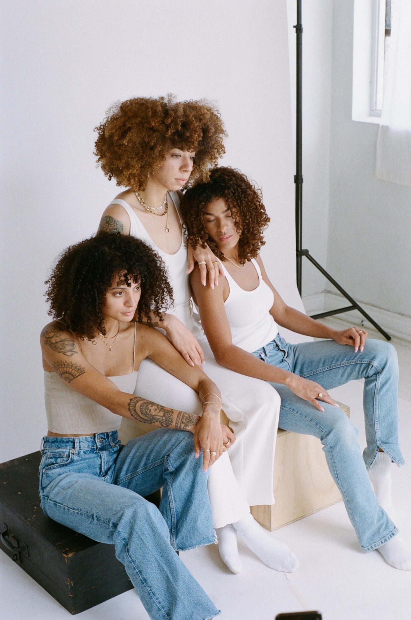 Three models with full, natural, frizzy curly hair in white tank tops and jeans leaning against each other against a white photo backdrop.