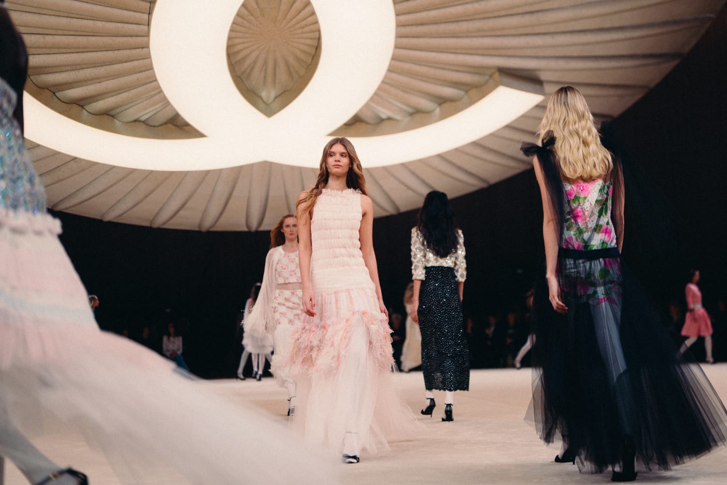 Chanel runway show. Large Chanel logo emblazoned on the ceiling with a group of models walking.