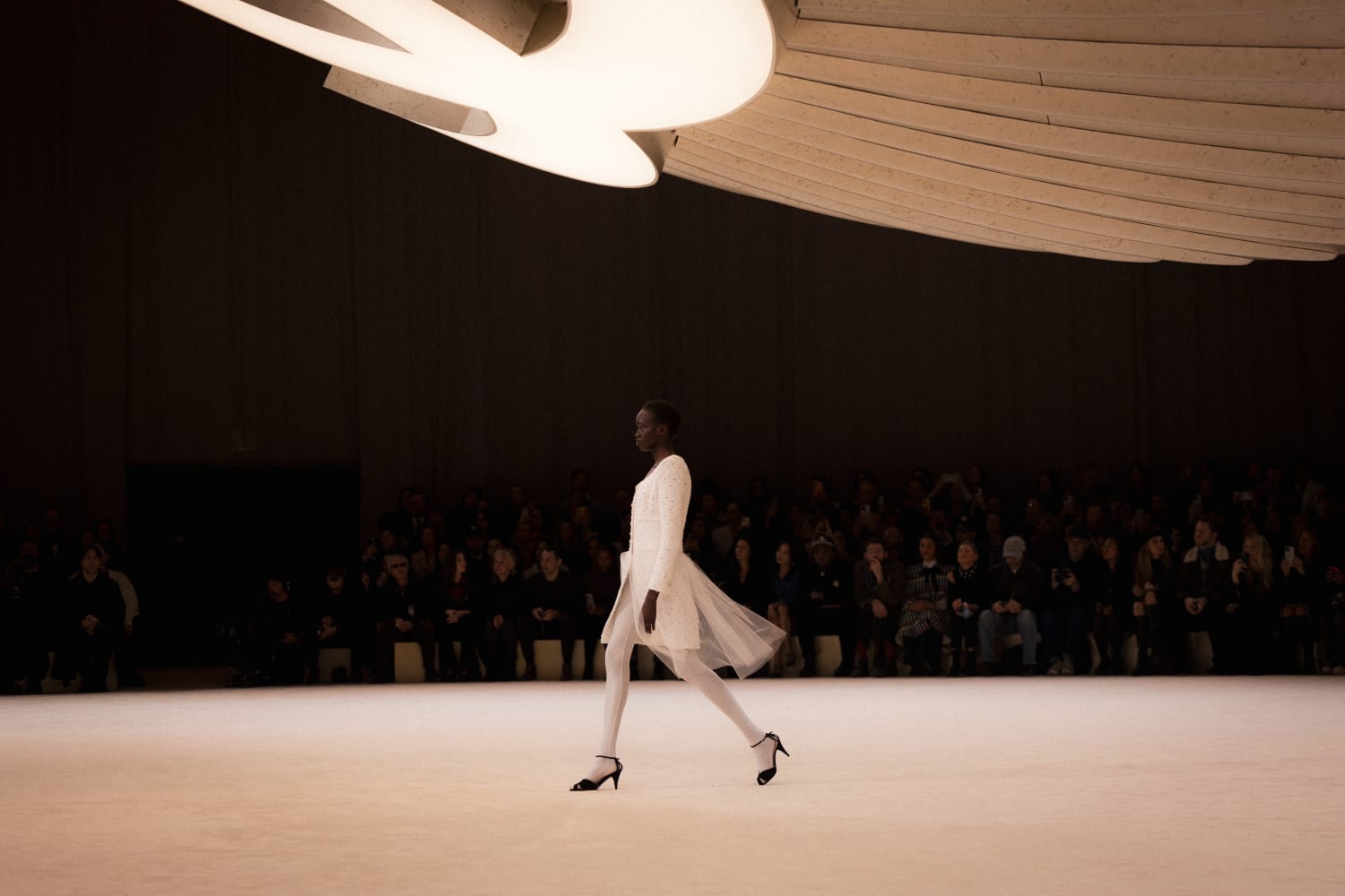 Chanel runway show. Large Chanel logo emblazoned on the ceiling with a model walking in a white gown.
