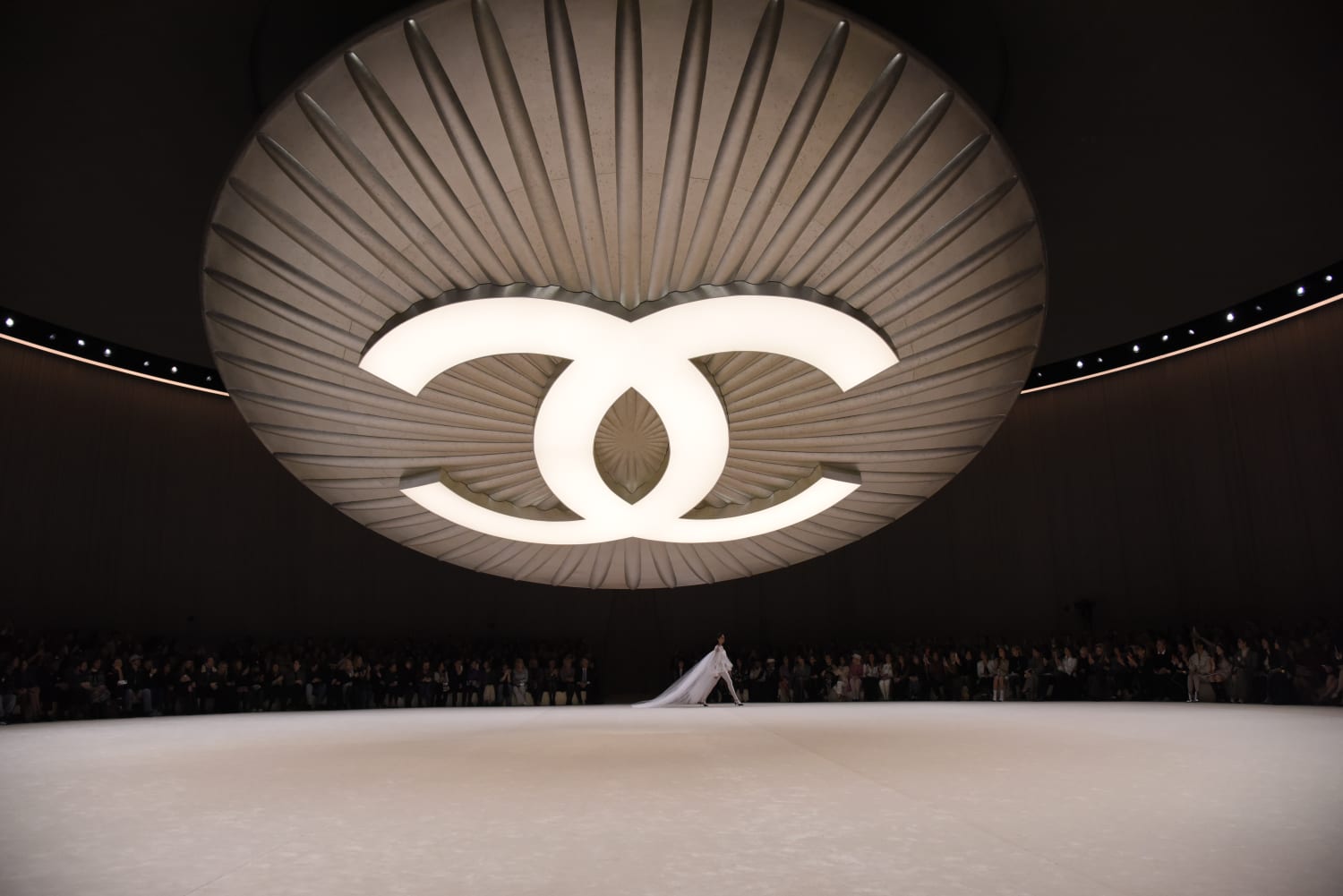 Chanel runway show. Large Chanel logo emblazoned on the ceiling with a model walking in a white gown.