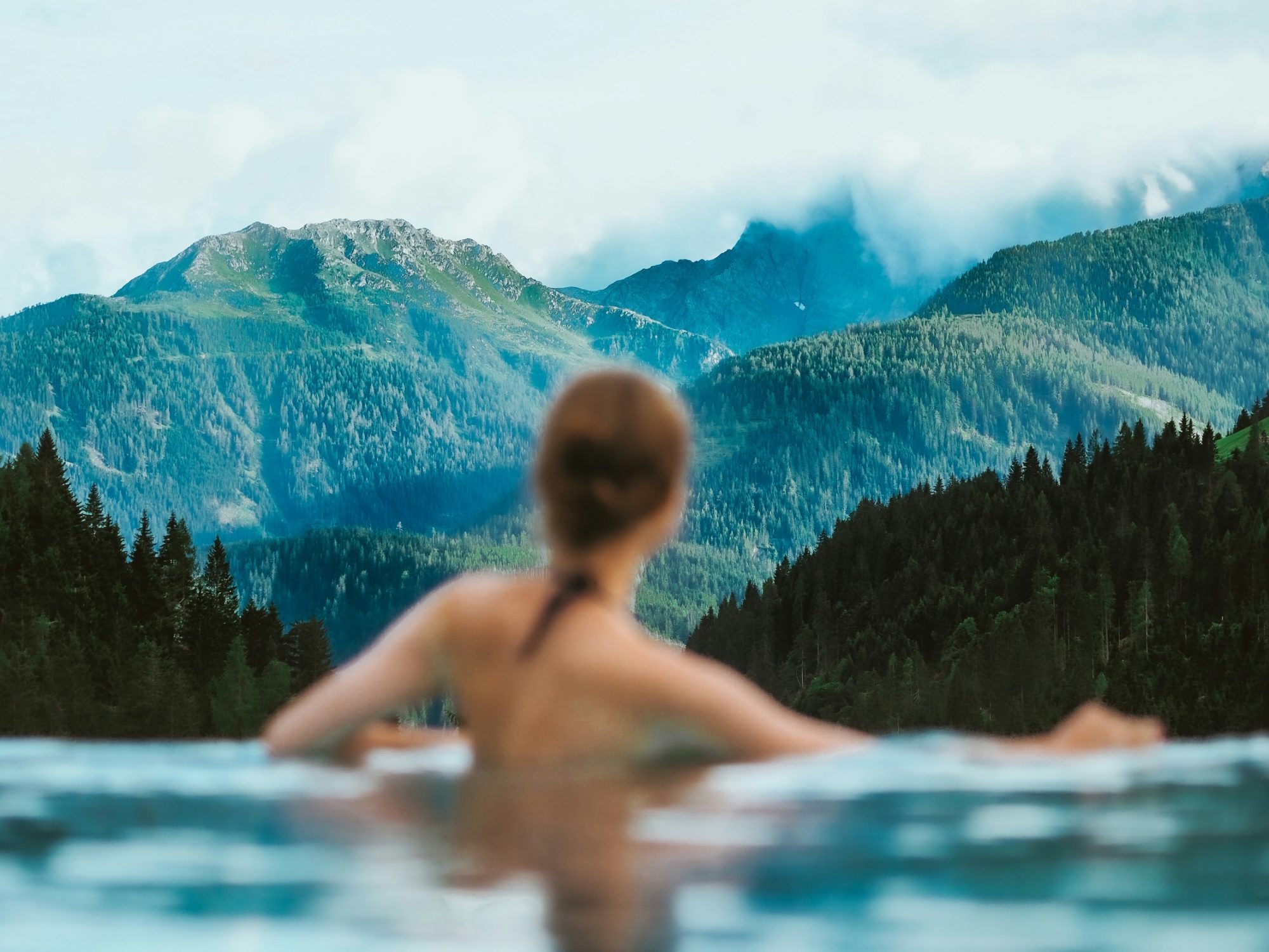 Woman sitting about waist-deep in an infinity pool overlooking a mountainous range with cloud cover.
