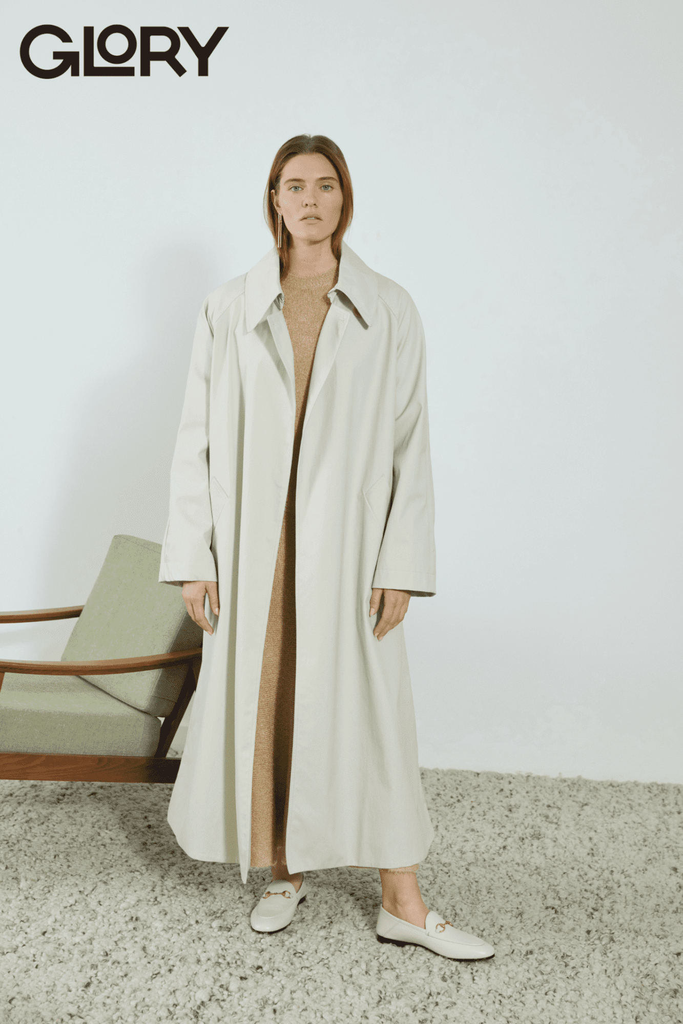 Alysha Newman standing up in a white trench coat wearing white shoes against a lounge chair and shag carpet.