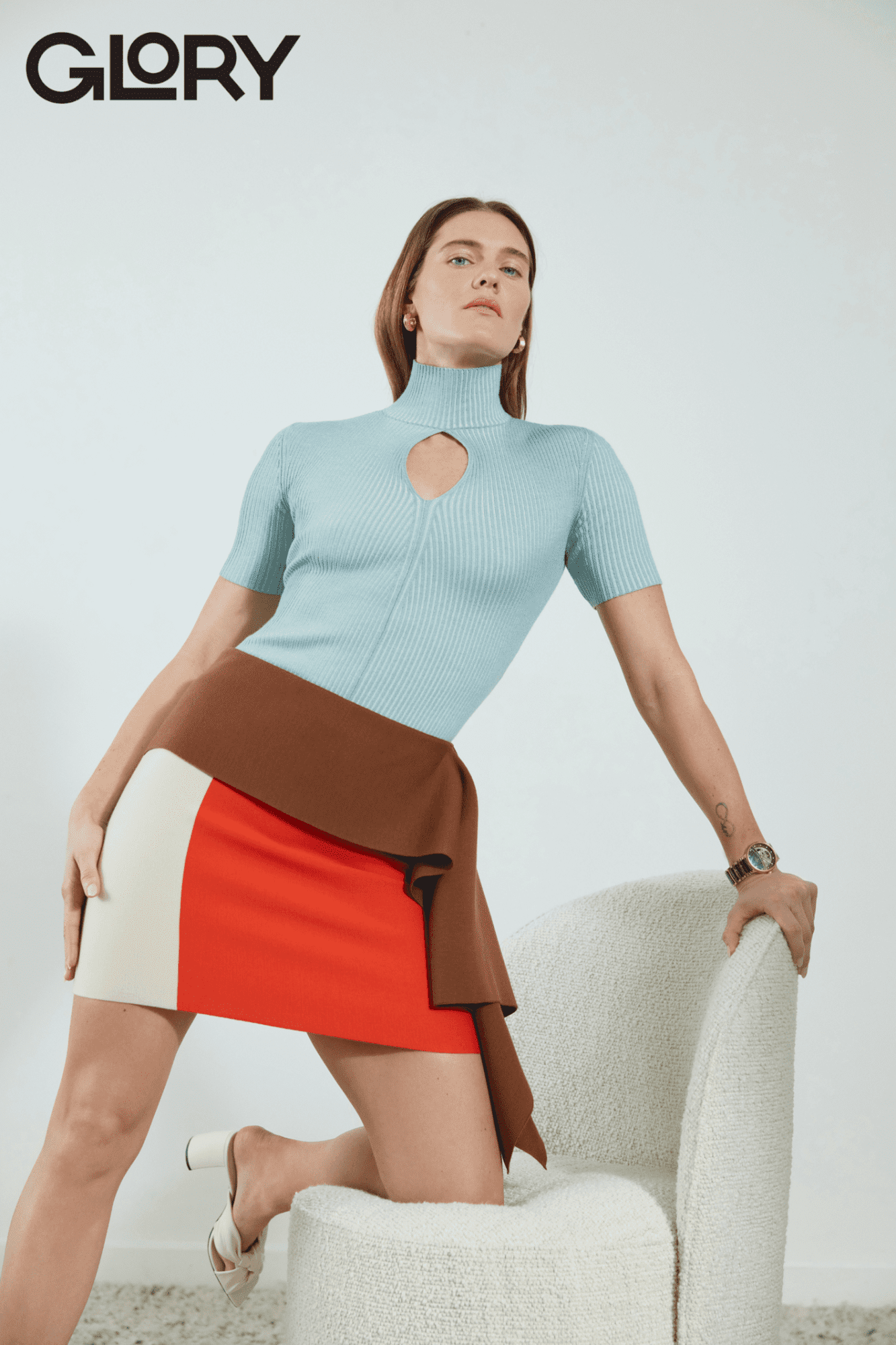 Alysha Newman kneeling confidently with one knee and an arm on a plush white lounge chair. She is wearing a light blue knitted top and colour blocked skirt with white high-heel shoes.