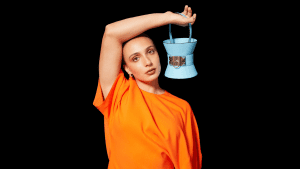 Marissa Papaconstantinou wearing an orange dress. Her arm is draped over her head and holding a light blue bucket purse.