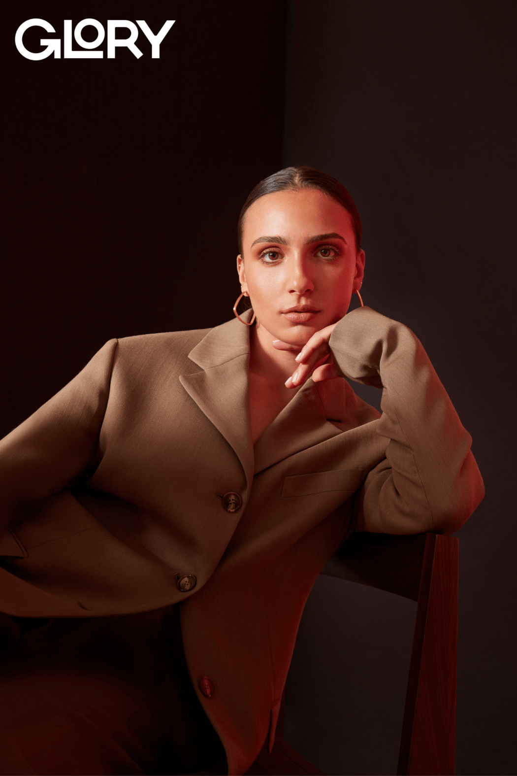 Marissa Papaconstantinou in a moody photo leaning against a chair wearing a brown suit jacket.