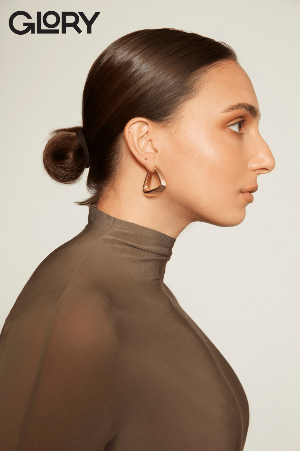 Side profile of Marissa Papaconstantinou wearing a sheer taupe dress. Her hair is tied in a tight bun and she is wearing gold earrings.