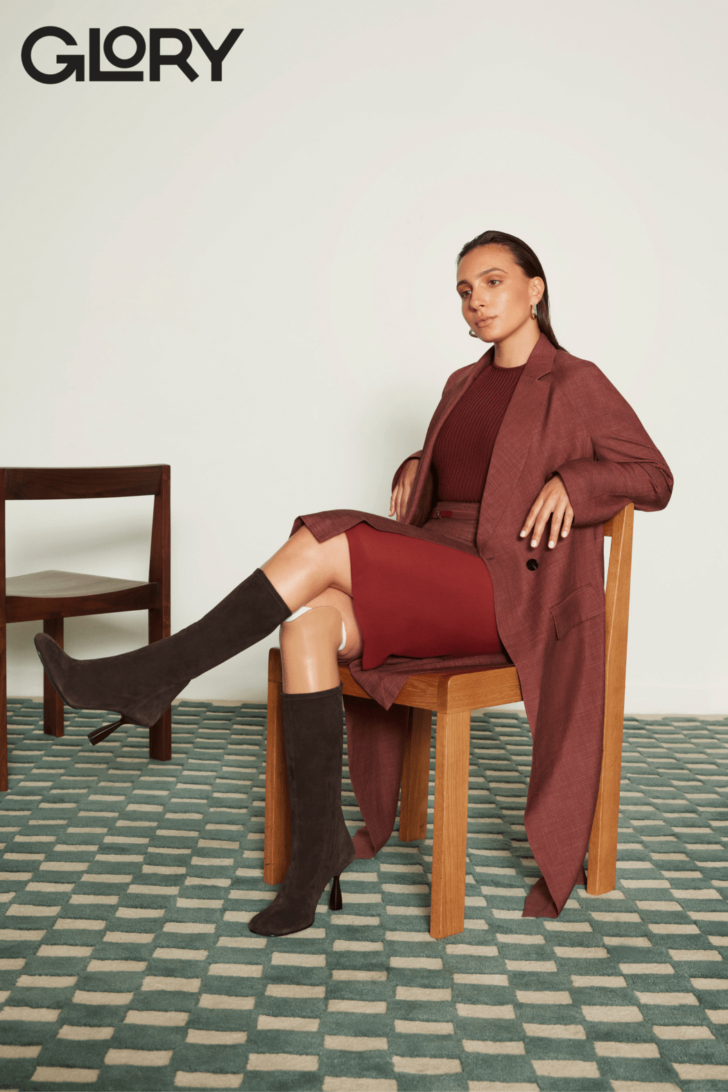 Marissa Papaconstantinou seated in a wooden chair on a checkered rug wearing a red jacket and skirt with tall brown boots.